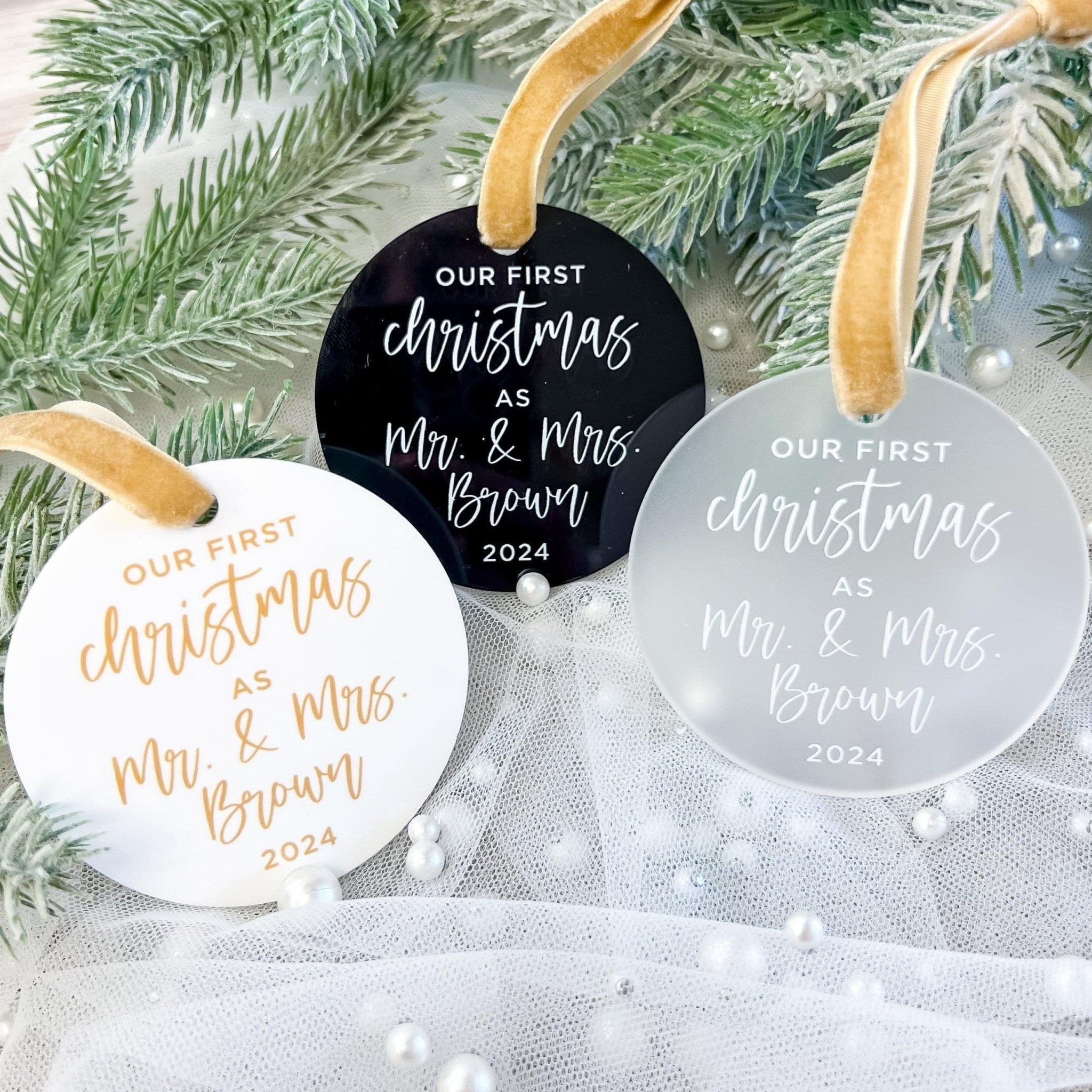 Three round acrylic ornaments in white, black, and clear with gold and silver text reading "Our First Christmas as Mr & Mrs Brown 2024" hanging on evergreen branches.