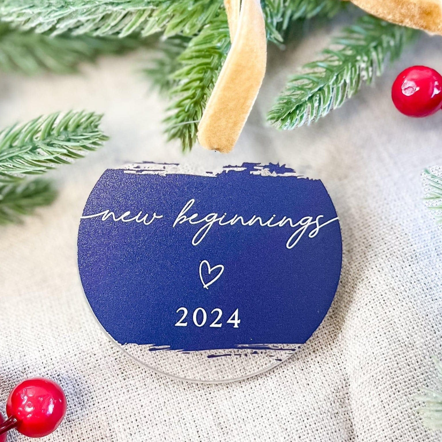 Navy blue acrylic ornament with "new beginnings" script text, small heart, and "2024" in gold lettering, hanging from tan ribbon among evergreen branches and red baubles.