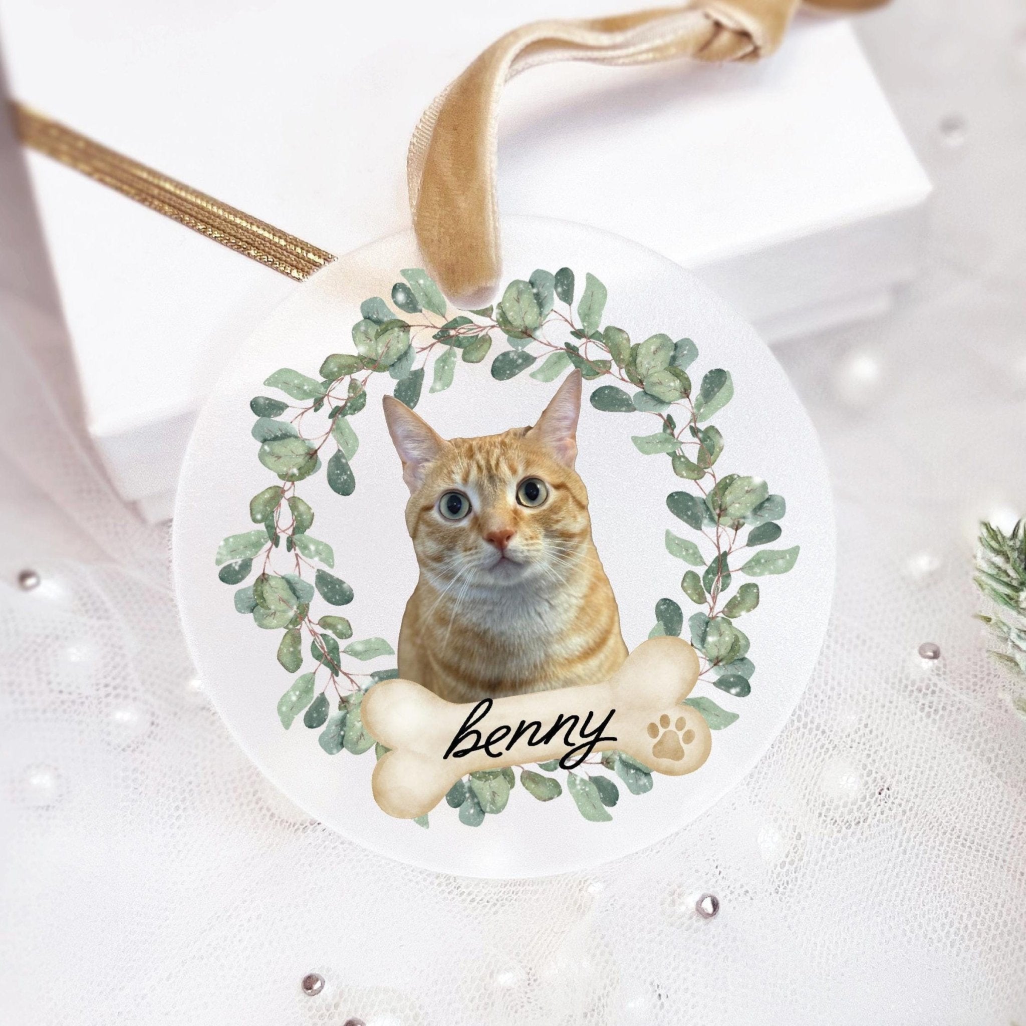White acrylic ornament with tabby cat photo surrounded by green eucalyptus wreath and personalized name "benny" in script, gold ribbon attachment.
