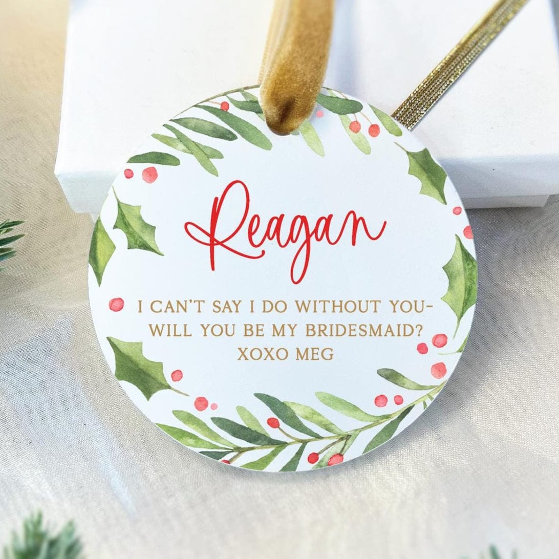 Round acrylic ornament with green foliage and pink berries border, personalized with "Reagan" in red script and bridesmaid proposal message in gold text.
