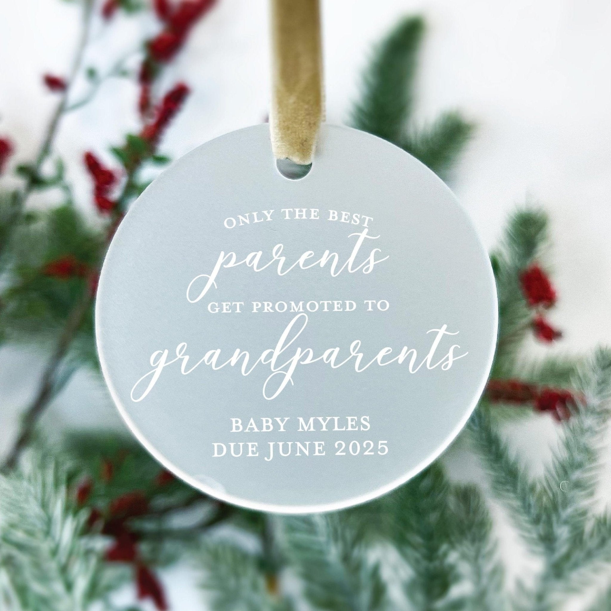Frosted acrylic round ornament with gold string, engraved with "Only the best parents get promoted to grandparents" and baby name and due date, displayed with evergreen and berry garland.