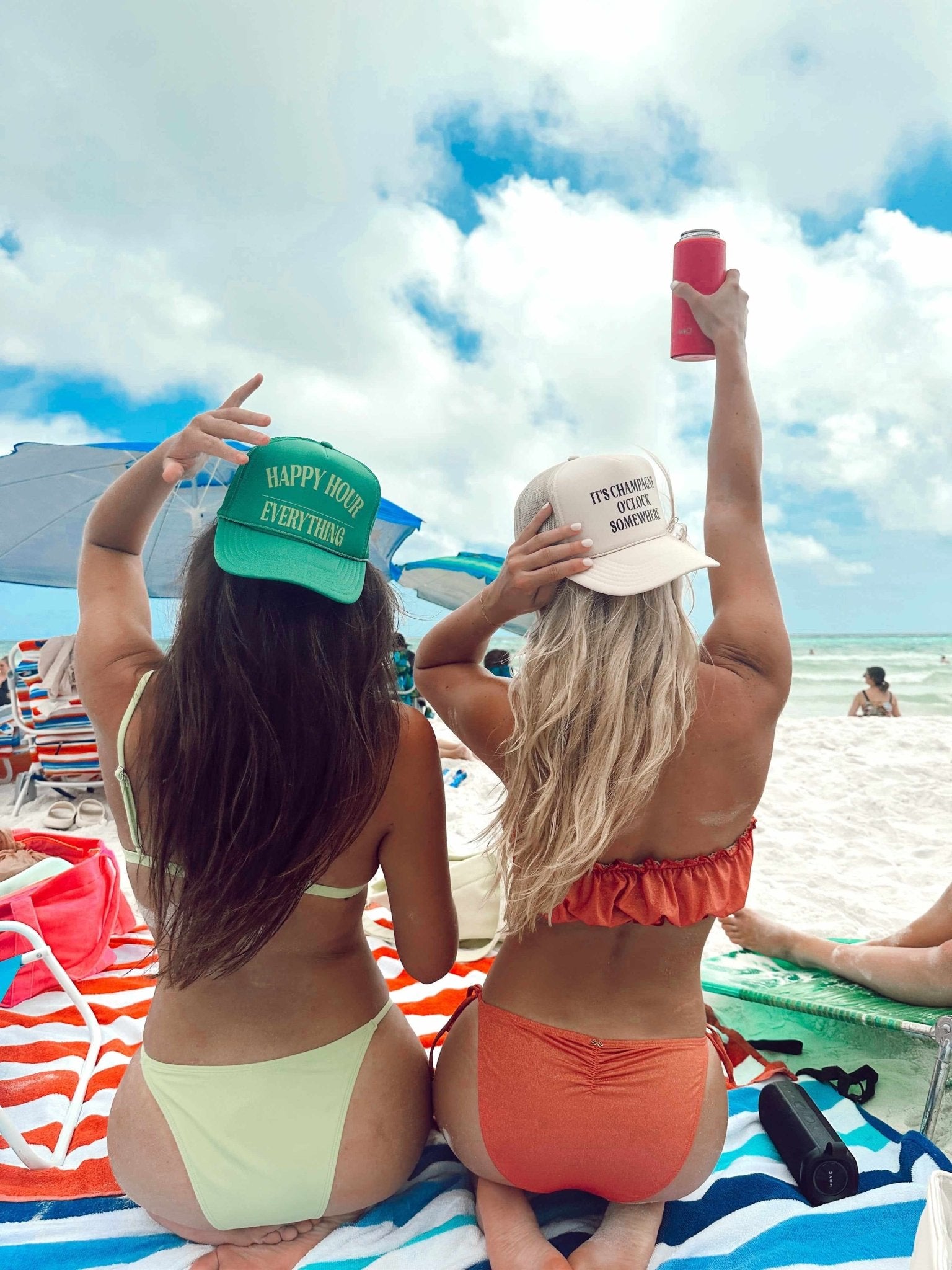 Two women on beach wearing personalized trucker hats, one green with "Happy Hour" text, one cream with custom design, holding pink cups toward sky