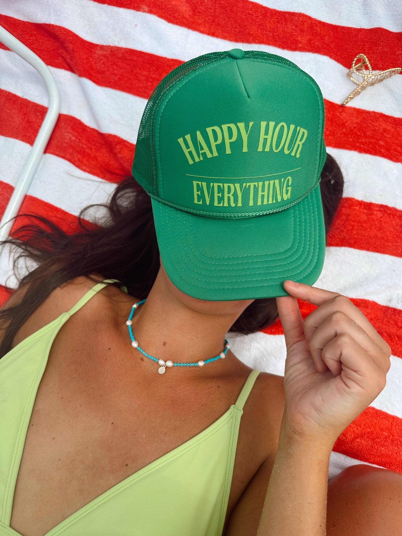 Green trucker hat with yellow "Happy Hour Everything" text, worn by person on red and white striped background.