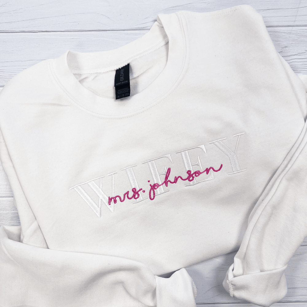 White crewneck sweatshirt with embroidered "mrs johnson" text in pink and white on the chest.