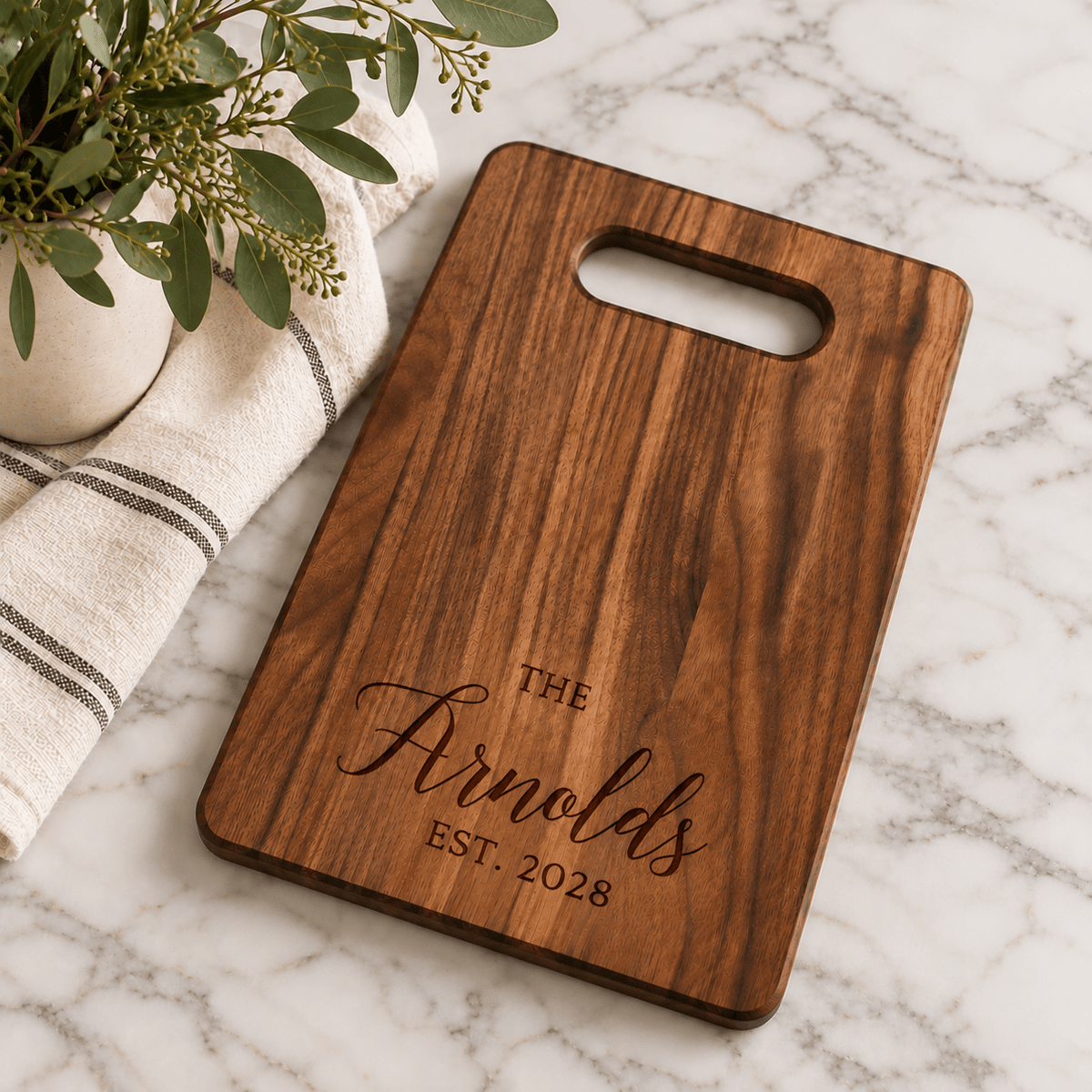 Custom walnut cutting board with engraved Arnold family name and established year 2028, styled on a marble countertop with greenery.