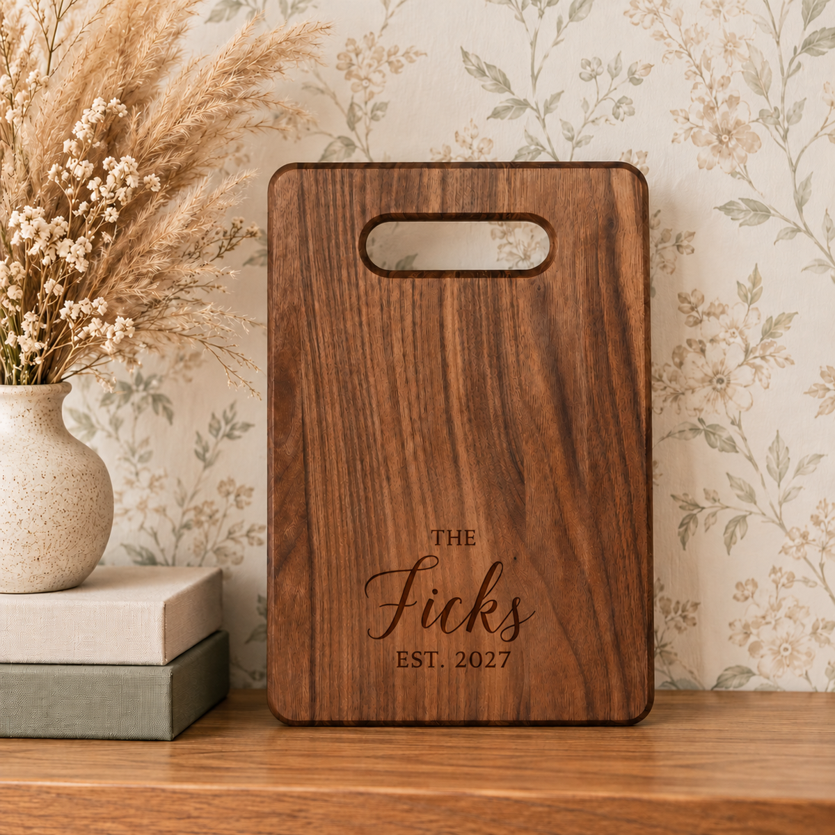 Engraved walnut cutting board featuring the Ficks family name and established year 2027, displayed upright against floral wallpaper.