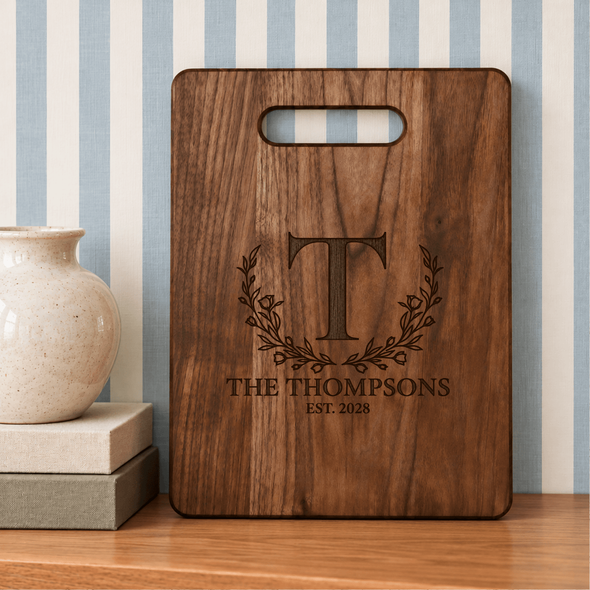 Custom walnut cutting board featuring Thompson family name, floral wreath monogram, and established year, displayed against blue and white striped wallpaper.
