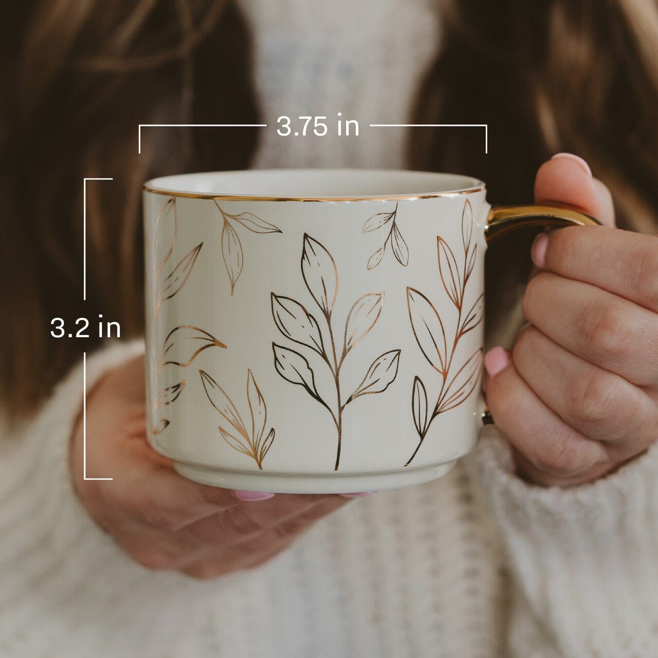 Cream ceramic mug with delicate botanical line drawings and gold rim, held in hands wearing a cream sweater