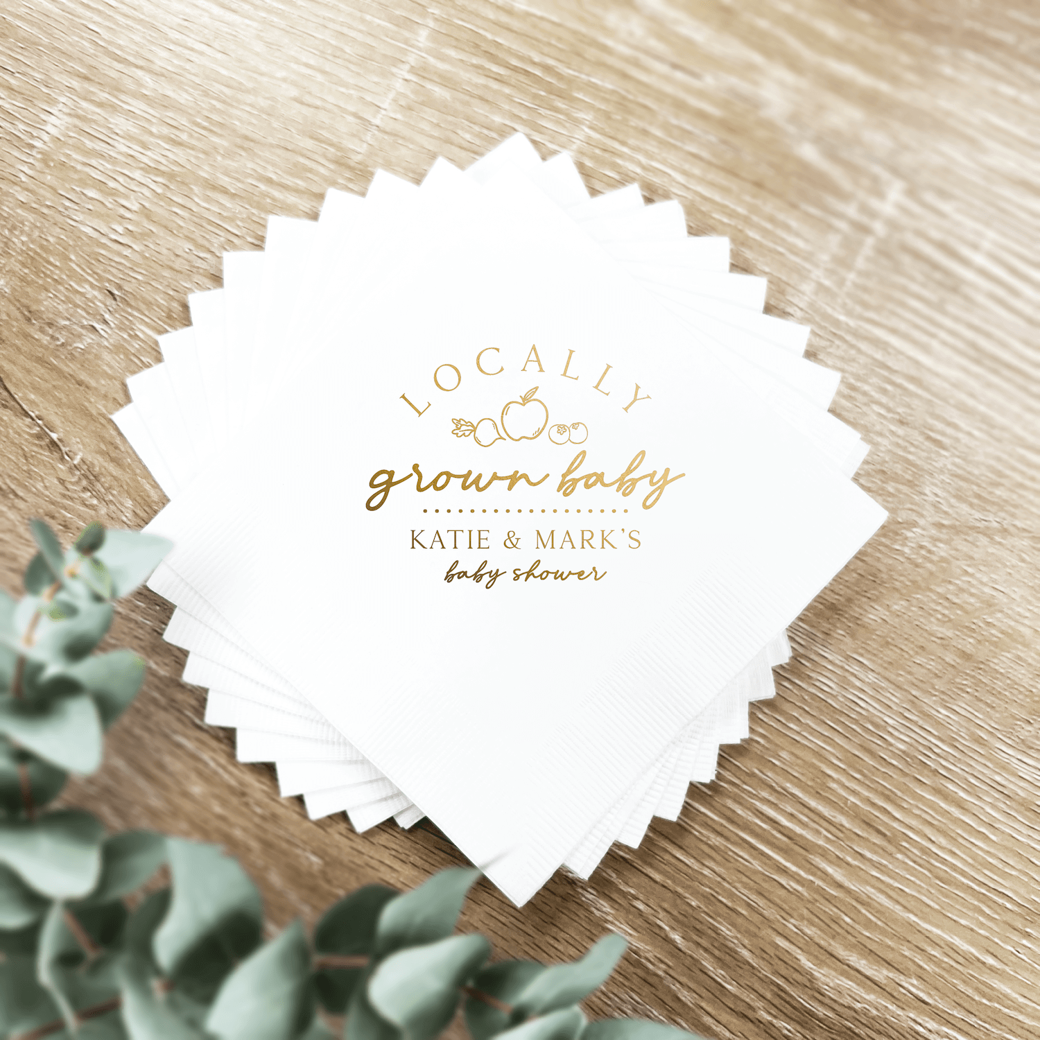 White napkins with scalloped edges and gold foil text reading "locally grown baby" and personalized names for a baby shower