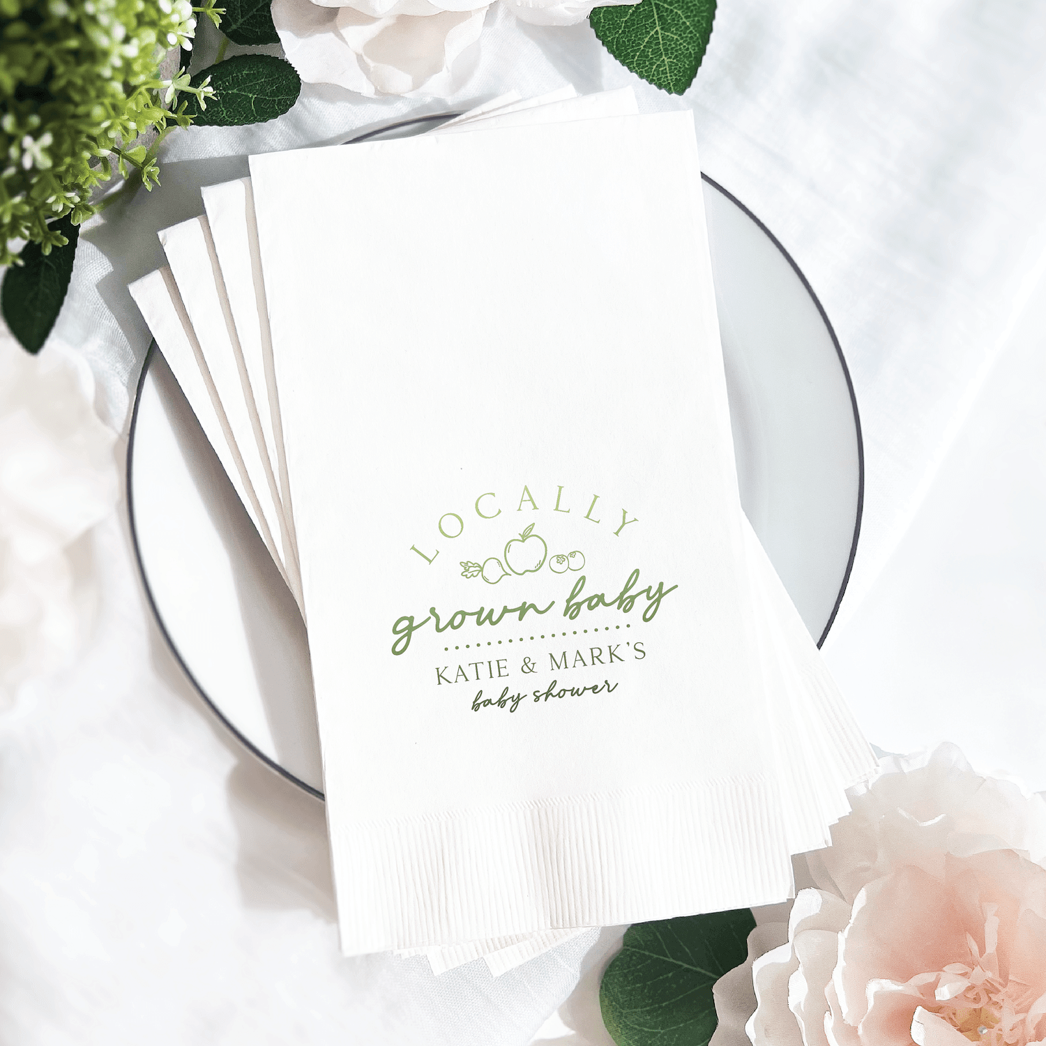 White napkins with gold foil stamped text reading "locally grown baby" and "Katie & Mark's baby shower" displayed on a place setting with flowers.