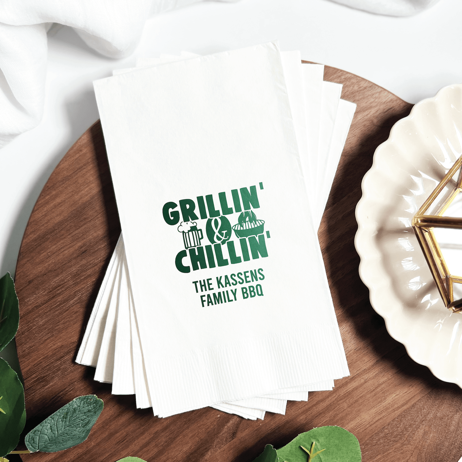 Stack of white napkins with green foil-stamped text reading "Grillin' & Chillin'" and "The Kassens Family BBQ" on wooden surface.