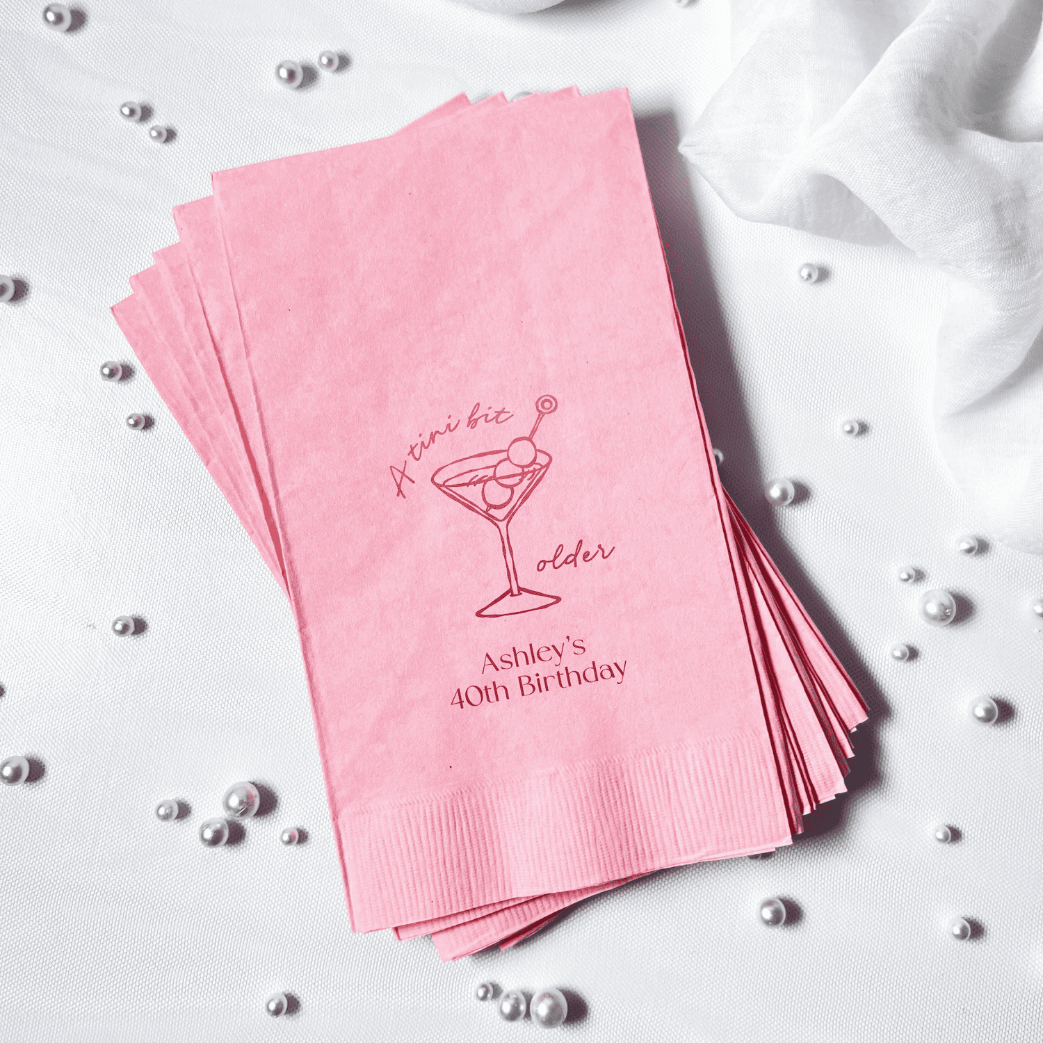 Stack of pink cocktail napkins with foil-stamped martini design and personalized names, surrounded by silver beads.