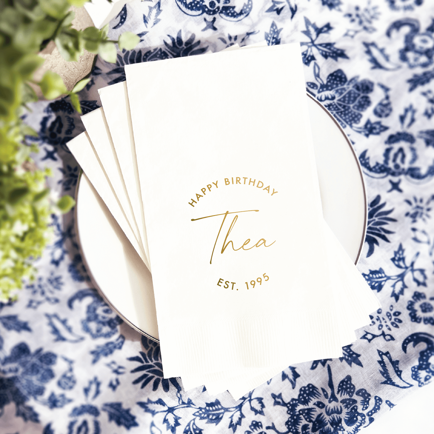 Stack of white napkins with gold foil "Happy Birthday Thea Est. 1993" personalization on blue floral plate