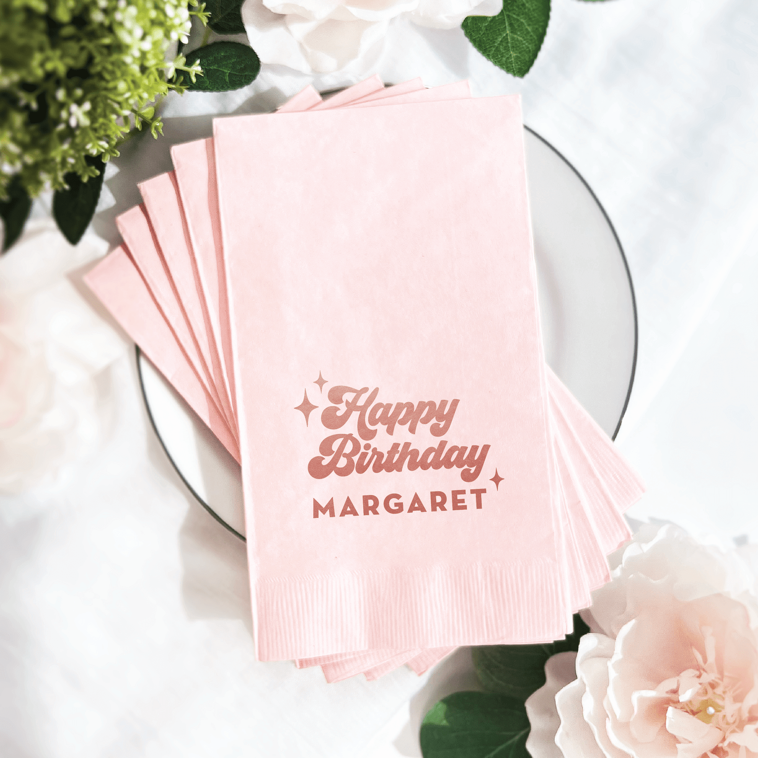 Stack of pink napkins with "Happy Birthday MARGARET" in rose gold foil stamping, displayed on white plate with flowers