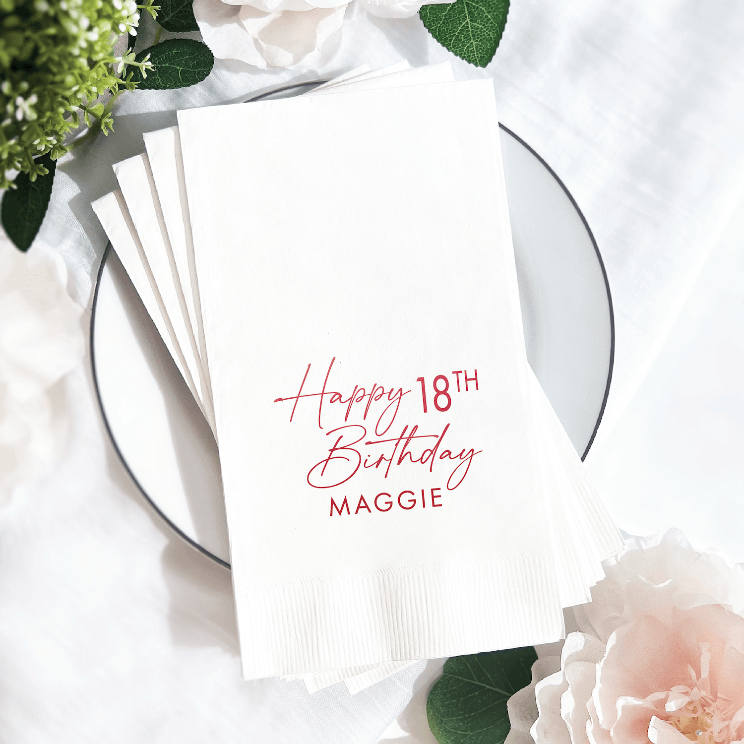 White napkin with pink foil-stamped text reading "Happy 18th Birthday Maggie" on a place setting with flowers