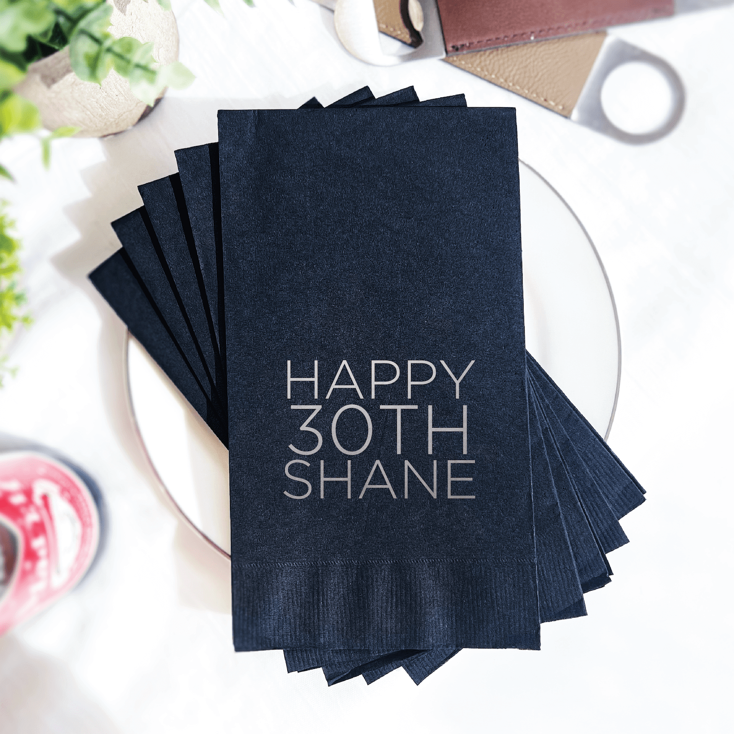 Stack of navy blue cocktail napkins with silver foil "Happy 30th Shane" text, displayed on a white plate.
