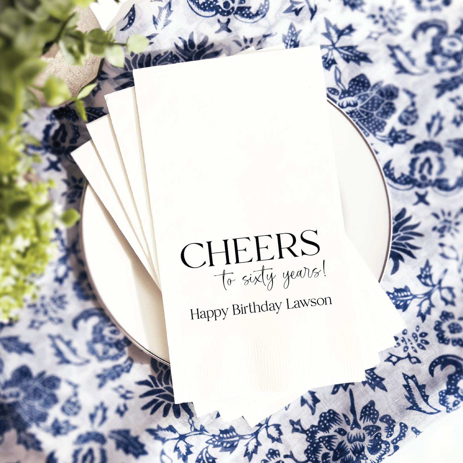 Stack of white napkins with "Cheers to sixty years" and "Happy Birthday Lawson" in black and silver foil stamping, displayed on blue and white patterned plate.