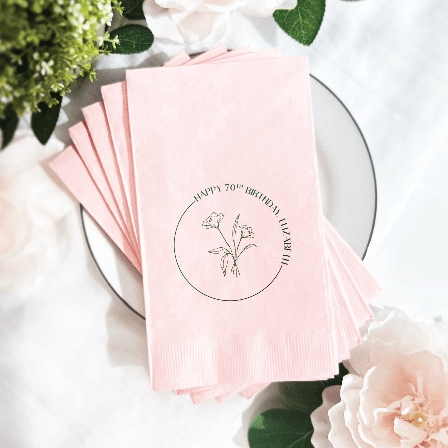 Stack of pink napkins with floral design and "Happy 70th Birthday" text in circular botanical illustration on white plate with flowers.