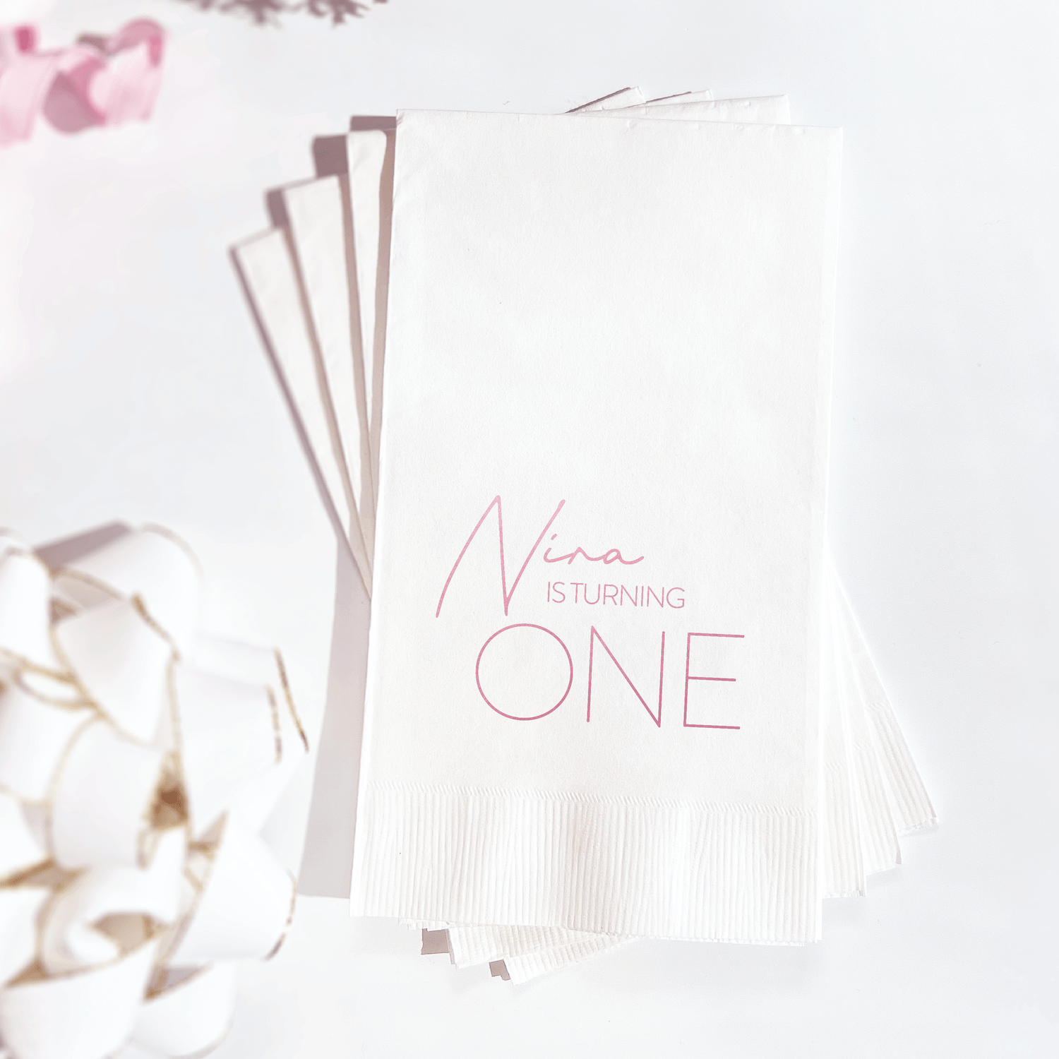 Stack of white napkins with pink foil-stamped text reading "Nina TURNING ONE" for a first birthday celebration.