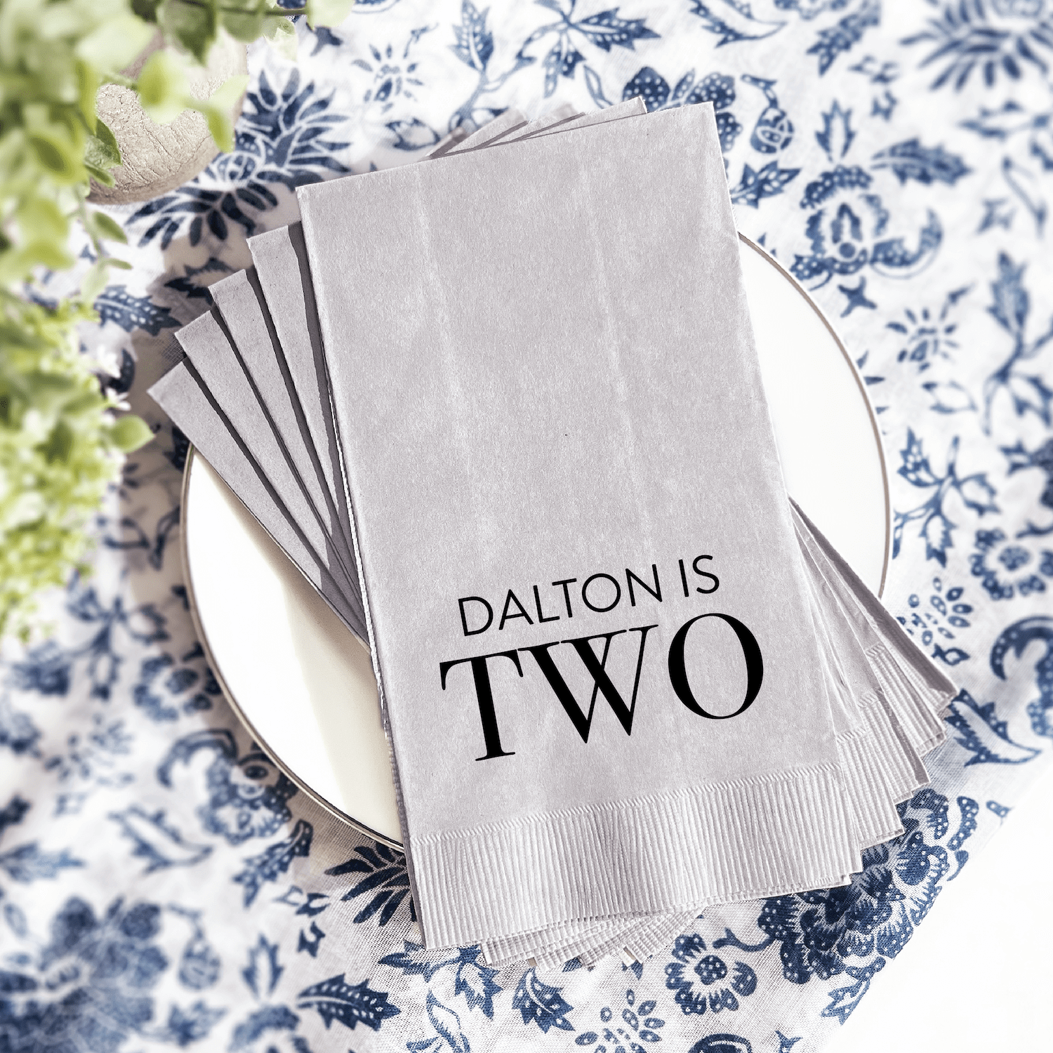 White napkin with "DALTON IS TWO" foil stamped text, displayed on plate with blue floral pattern background.
