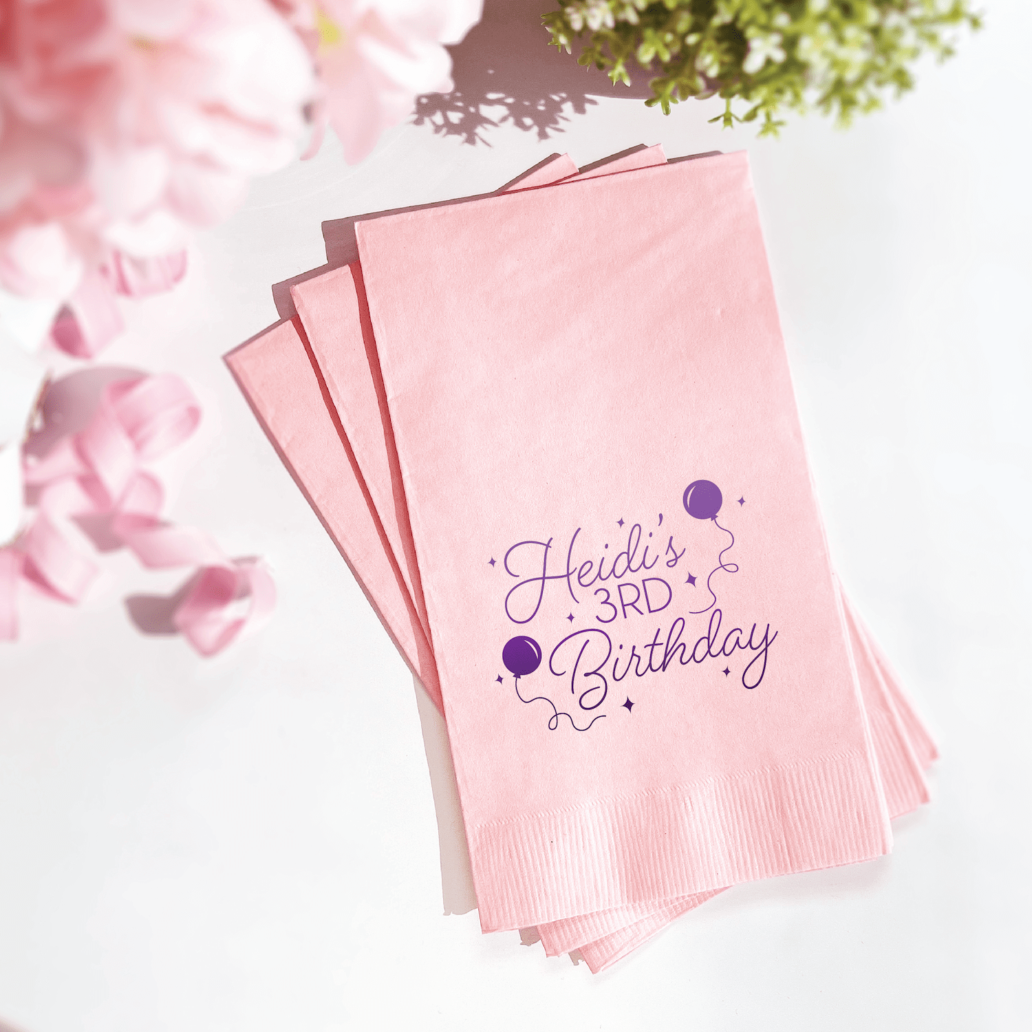 Stack of light pink napkins with purple foil-stamped text reading "Heidi's 3RD Birthday" and confetti balloon design