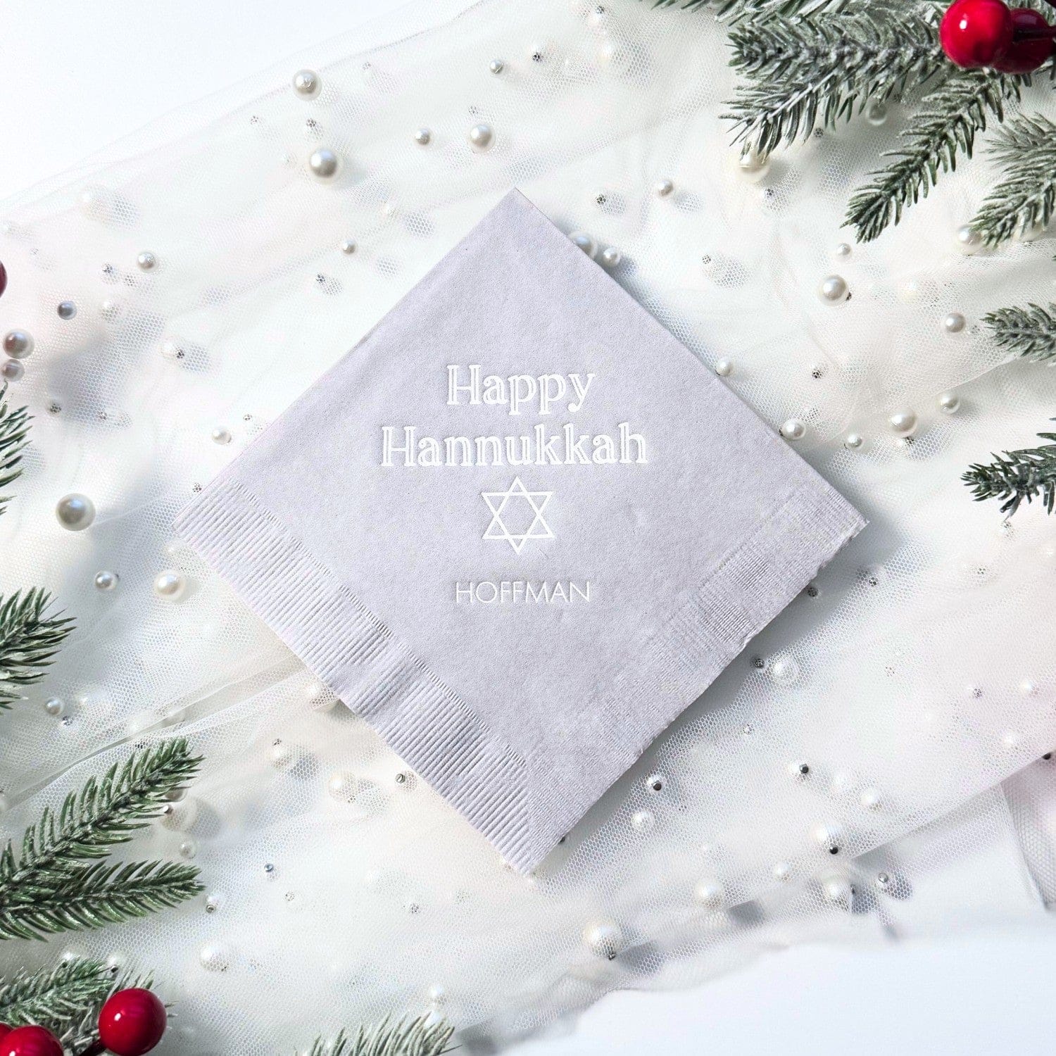 A light grey personalized paper napkin with a subtle embossed design is elegantly presented on a white sheer fabric adorned with pearls and frosted evergreen branches, suitable for a holiday celebration.