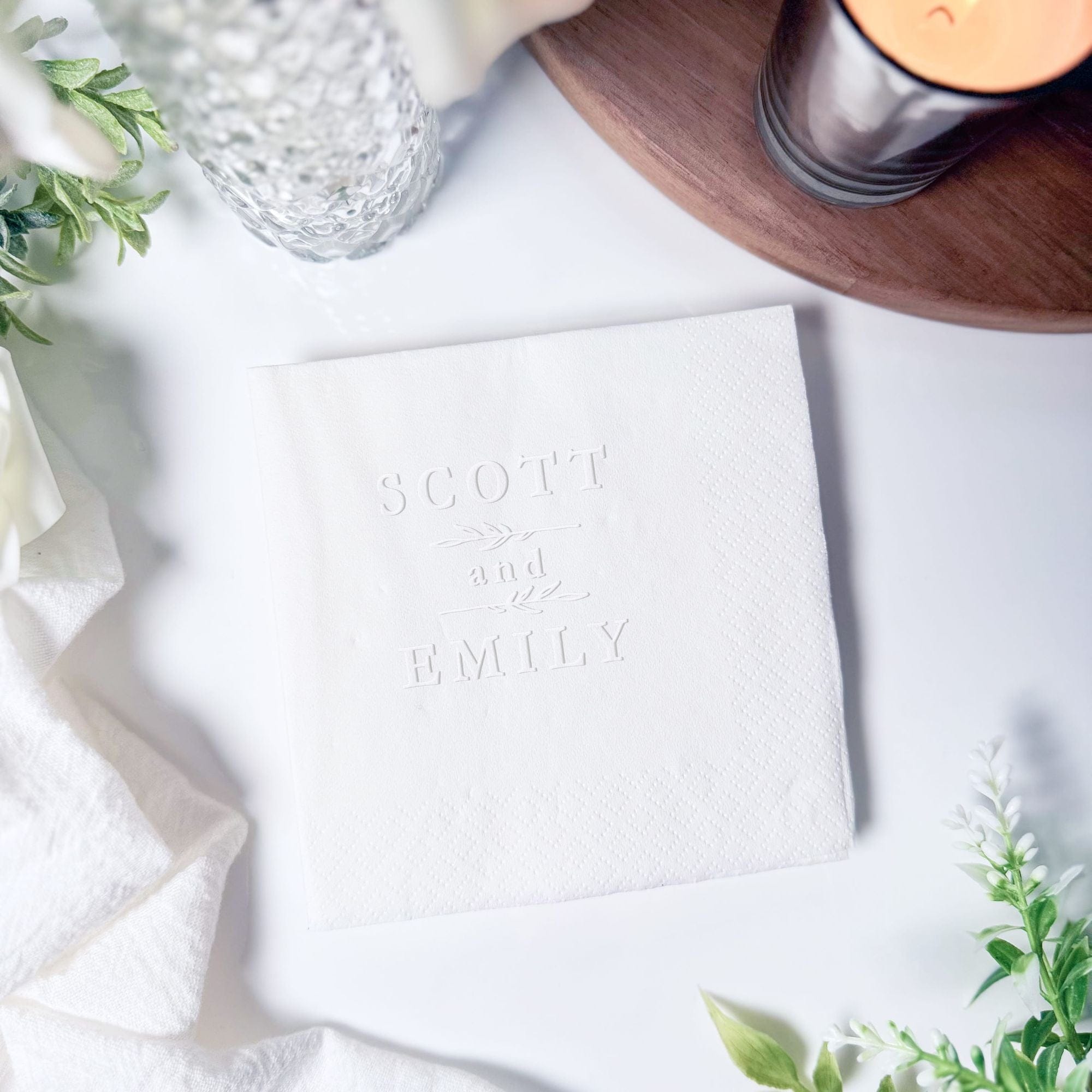 White embossed cocktail napkins with "Scott and Emily" names, surrounded by greenery and glassware on white surface.