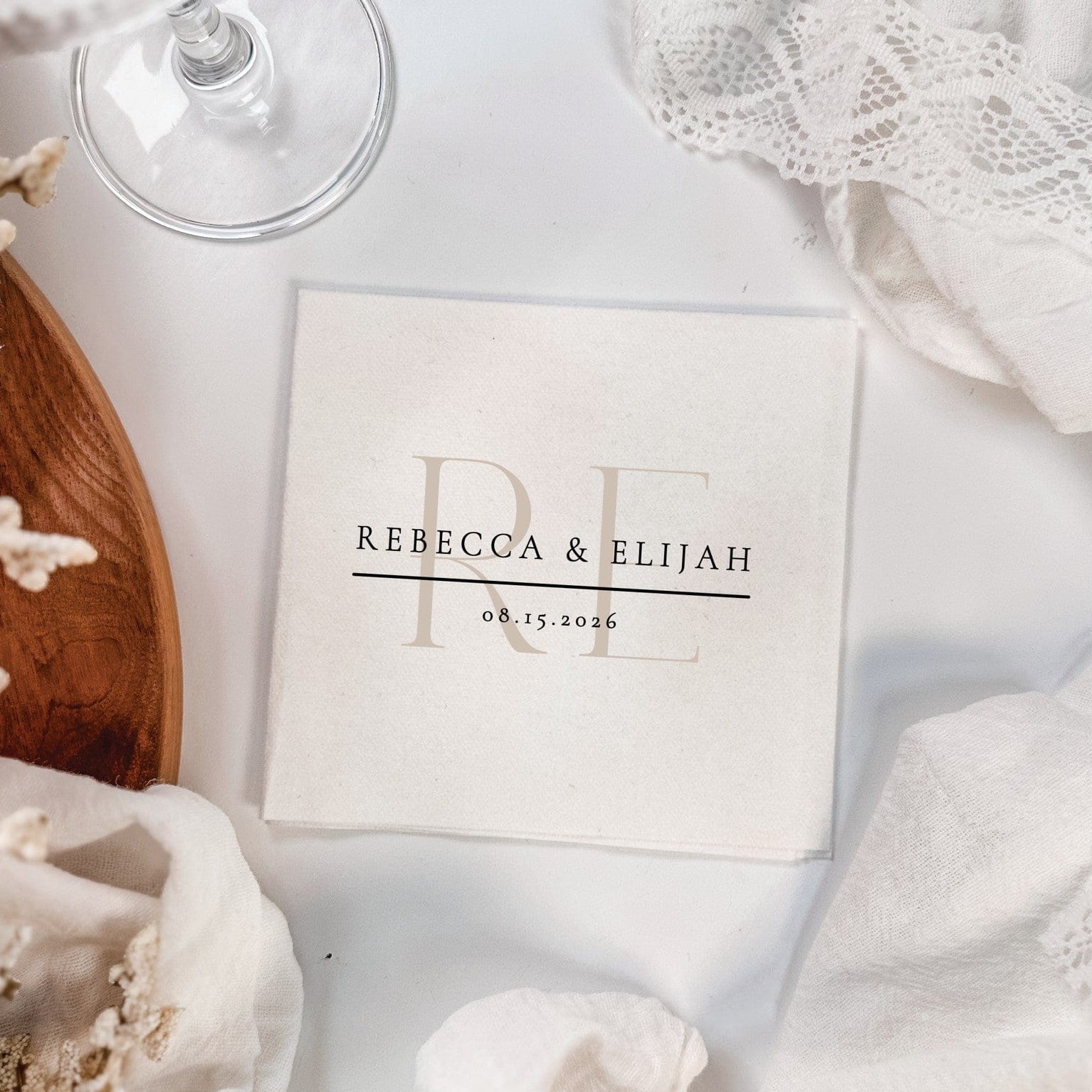 An off-white square napkin, crafted from a soft, absorbent material, displays large, overlapping initial designs alongside additional personalized lines of text, suitable for elegant wedding event decor.