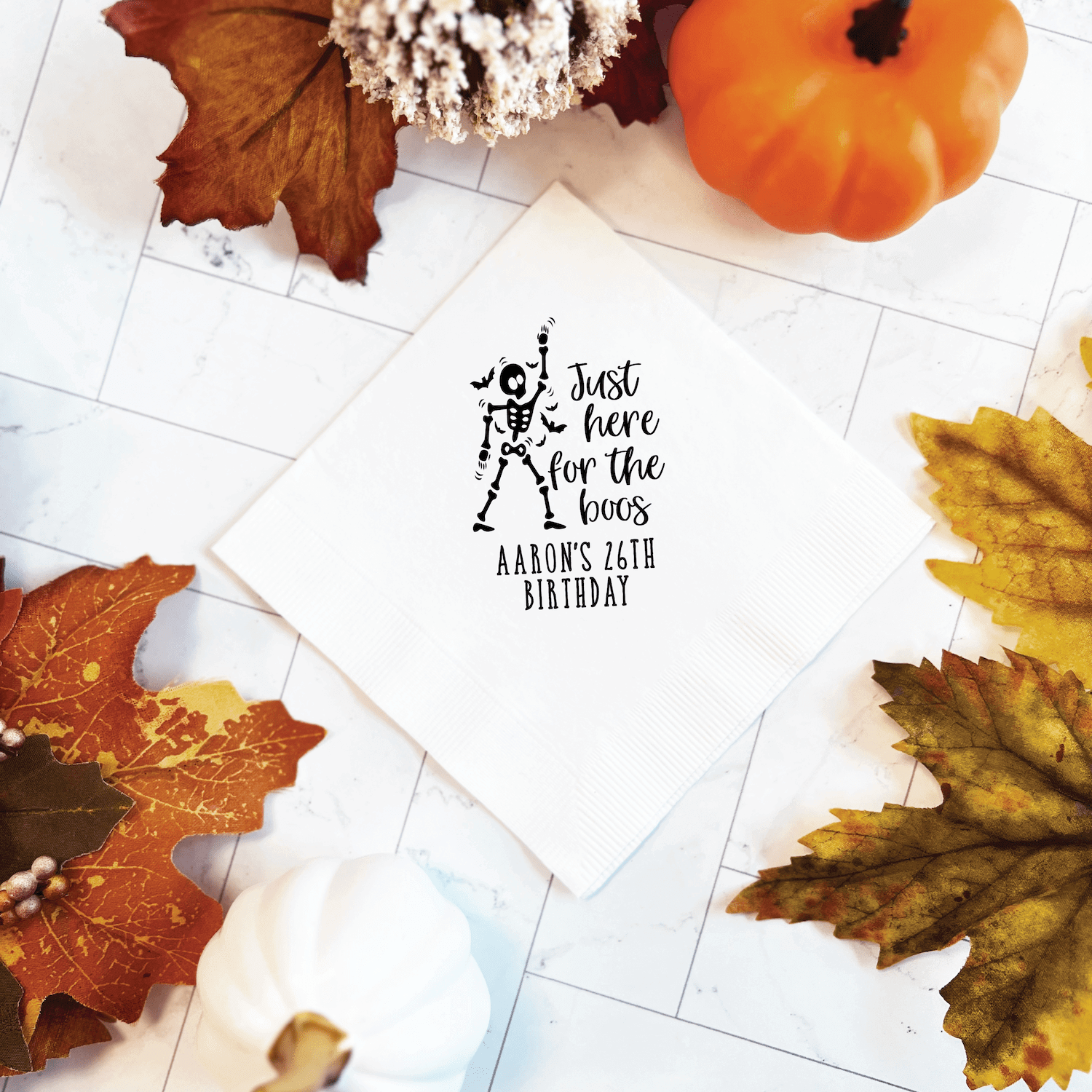 White napkin with skeleton design and "Just here for the boos" text personalized with Aaron's 26th Birthday in black foil stamping, surrounded by fall leaves and pumpkin decor.
