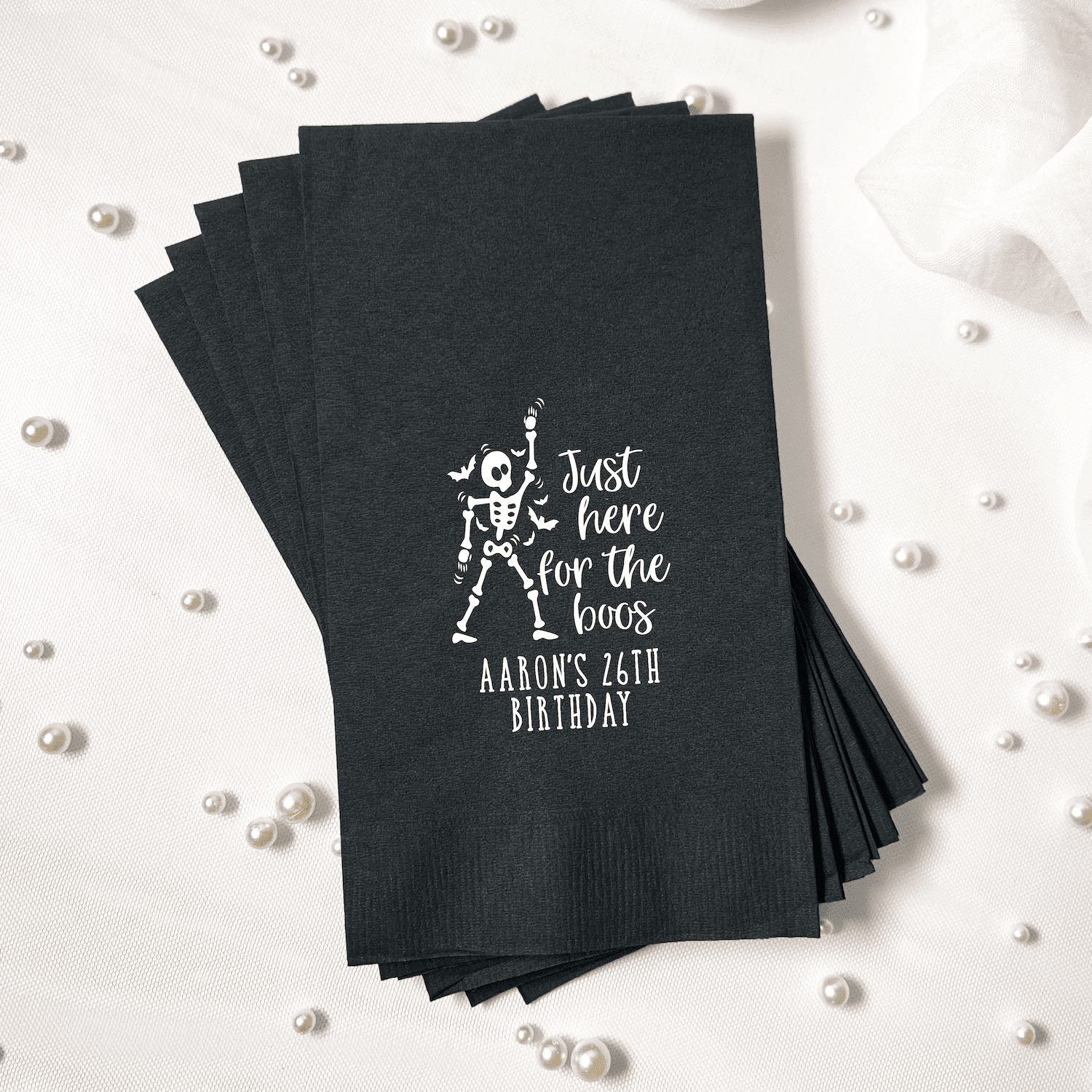 Stack of black cocktail napkins with white foil-stamped skeleton design and "Just Here for the Boos" text with personalized birthday date.