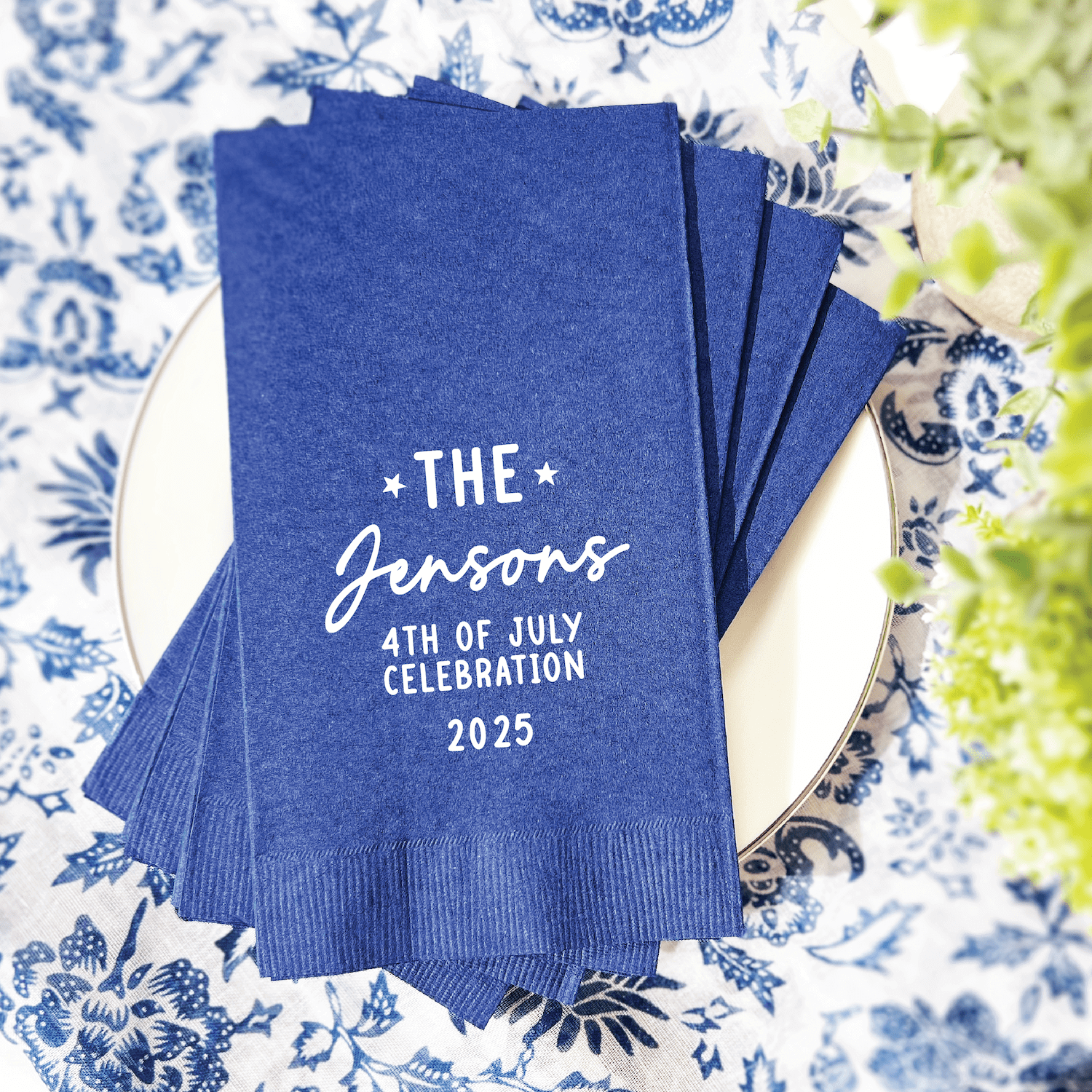 Blue cocktail napkins personalized with "The Jensons 4th of July Celebration 2025" in white foil lettering, displayed on a decorative plate.