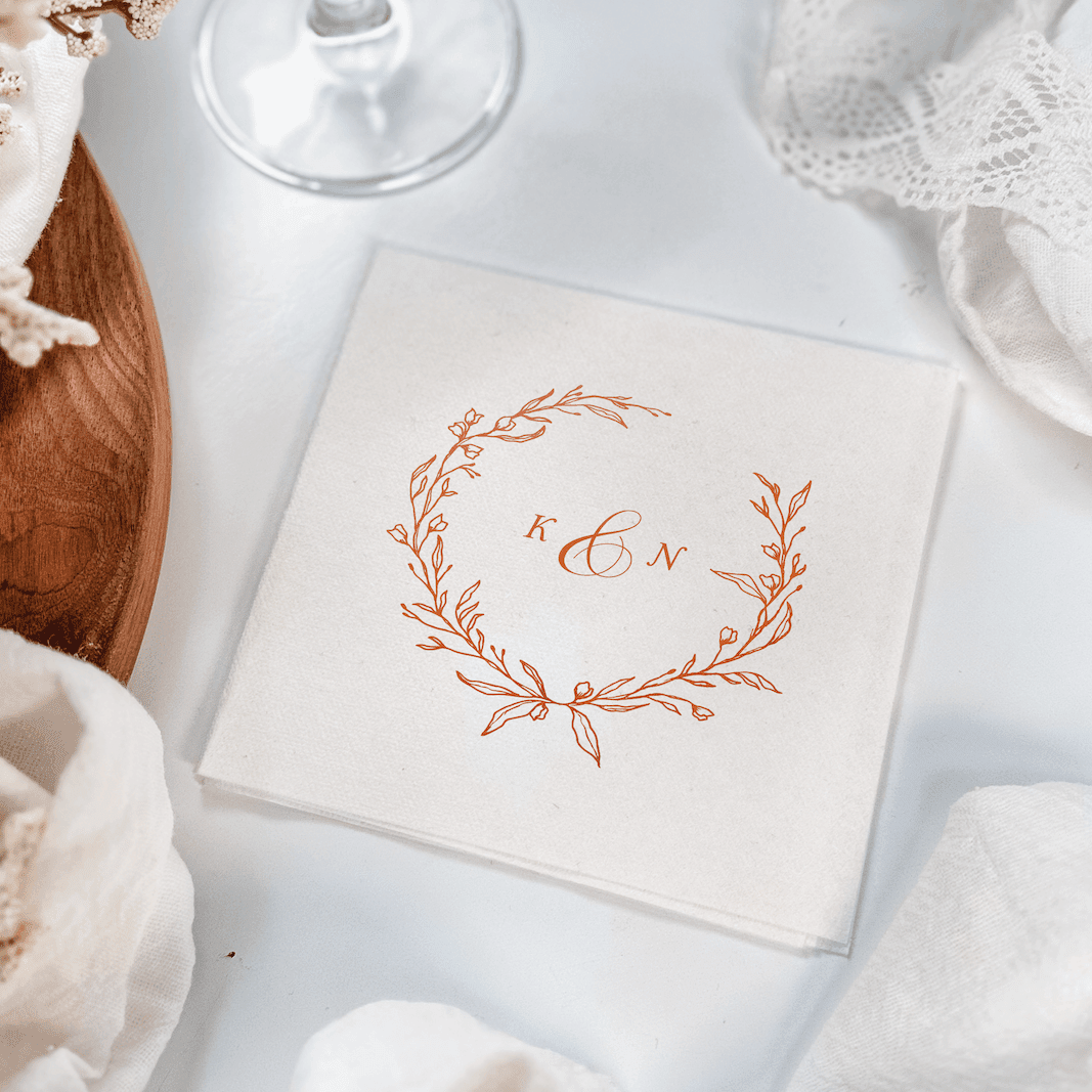 White cocktail napkin with copper foil stamped wreath design and monogram letter "E" in the center.