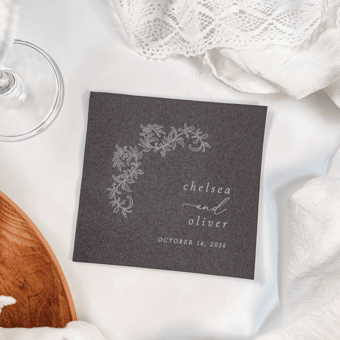 Dark gray cocktail napkin with silver floral crest design and personalized names "chelsea oliver" with wedding date October 14, 2024.