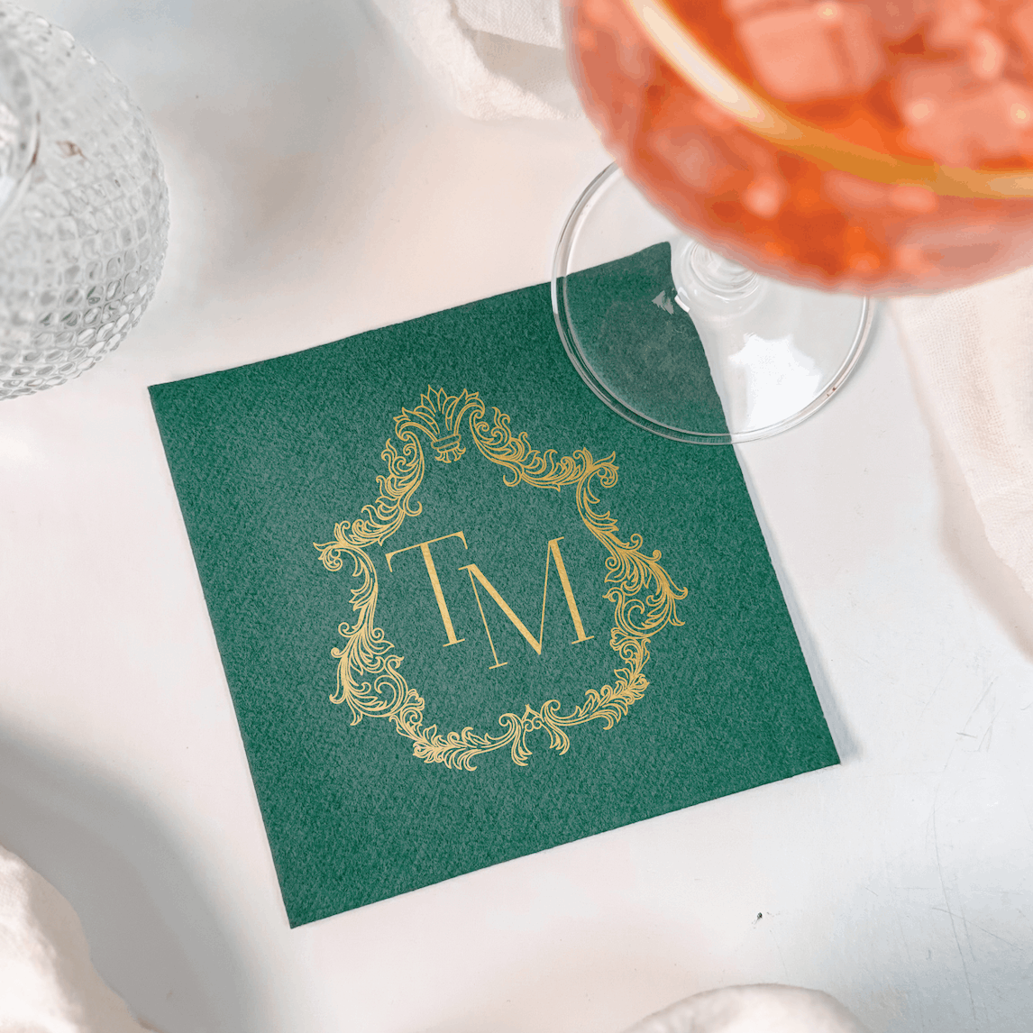 Green cocktail napkin with gold foil ornate frame monogram design and initials, styled with wine glass and decorative accents.