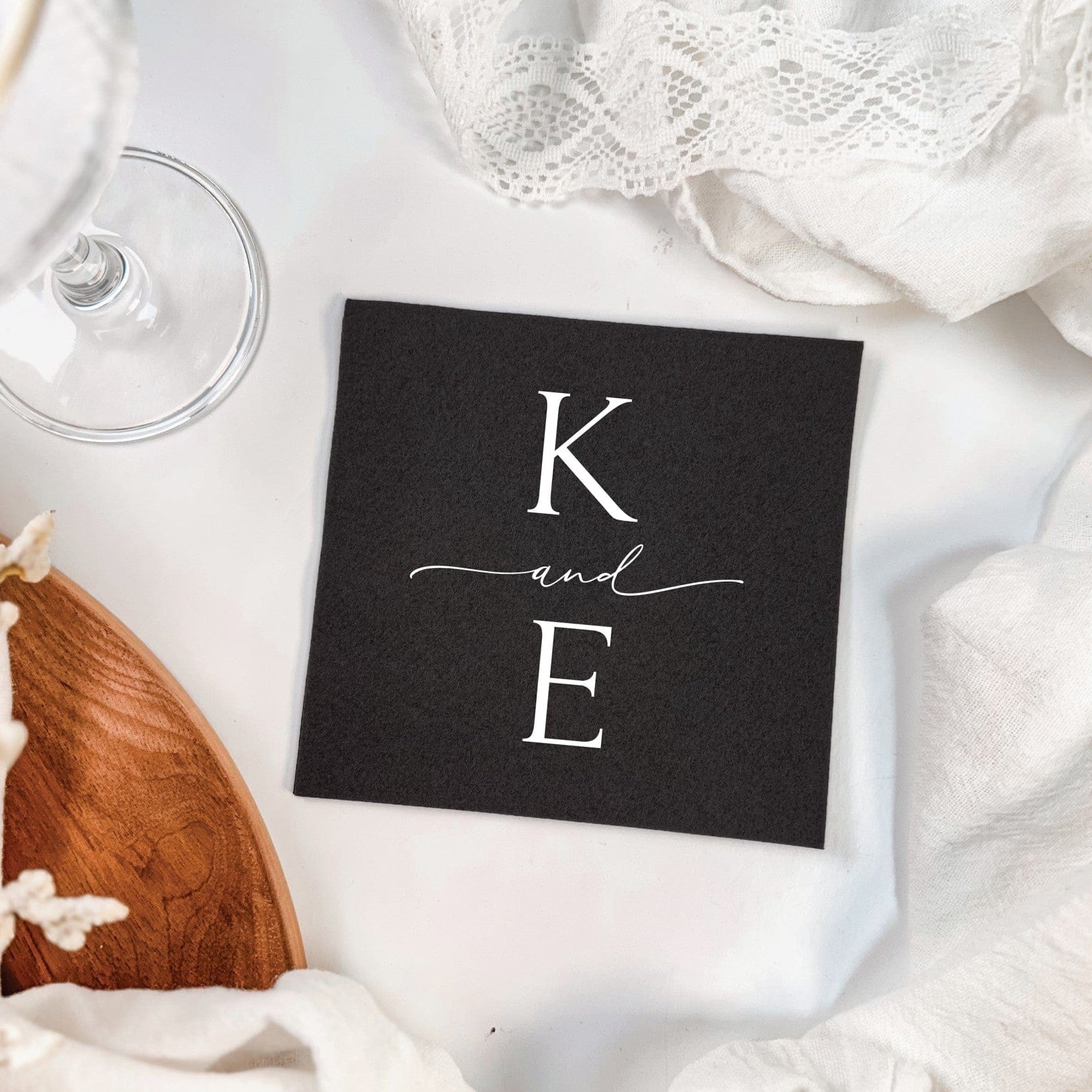 A black square personalized cocktail napkin featuring a custom white initial design, presented on a white surface with delicate lace and a clear wine glass, suitable for wedding or event decor.