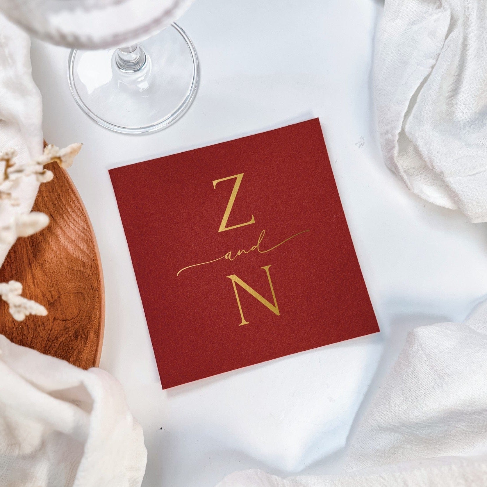 A square deep red paper napkin with elegant gold foil monogramming, styled with a glass and natural decor for a personalized wedding or event setting.