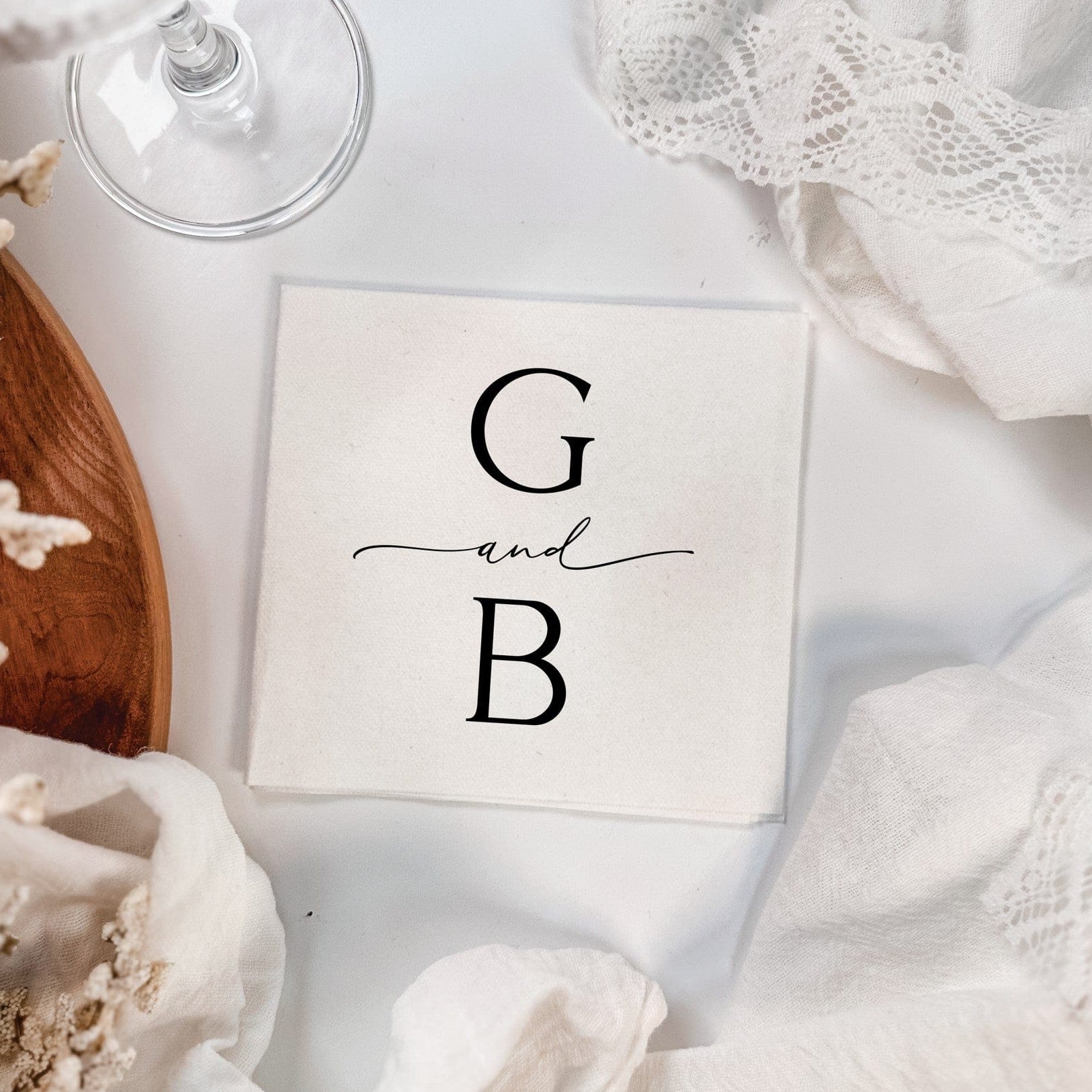 A personalized white linen-textured beverage napkin or coaster, customized with initial-style lettering, presented elegantly with draped white fabric, dried floral elements, and glassware, suitable for a wedding or special event.