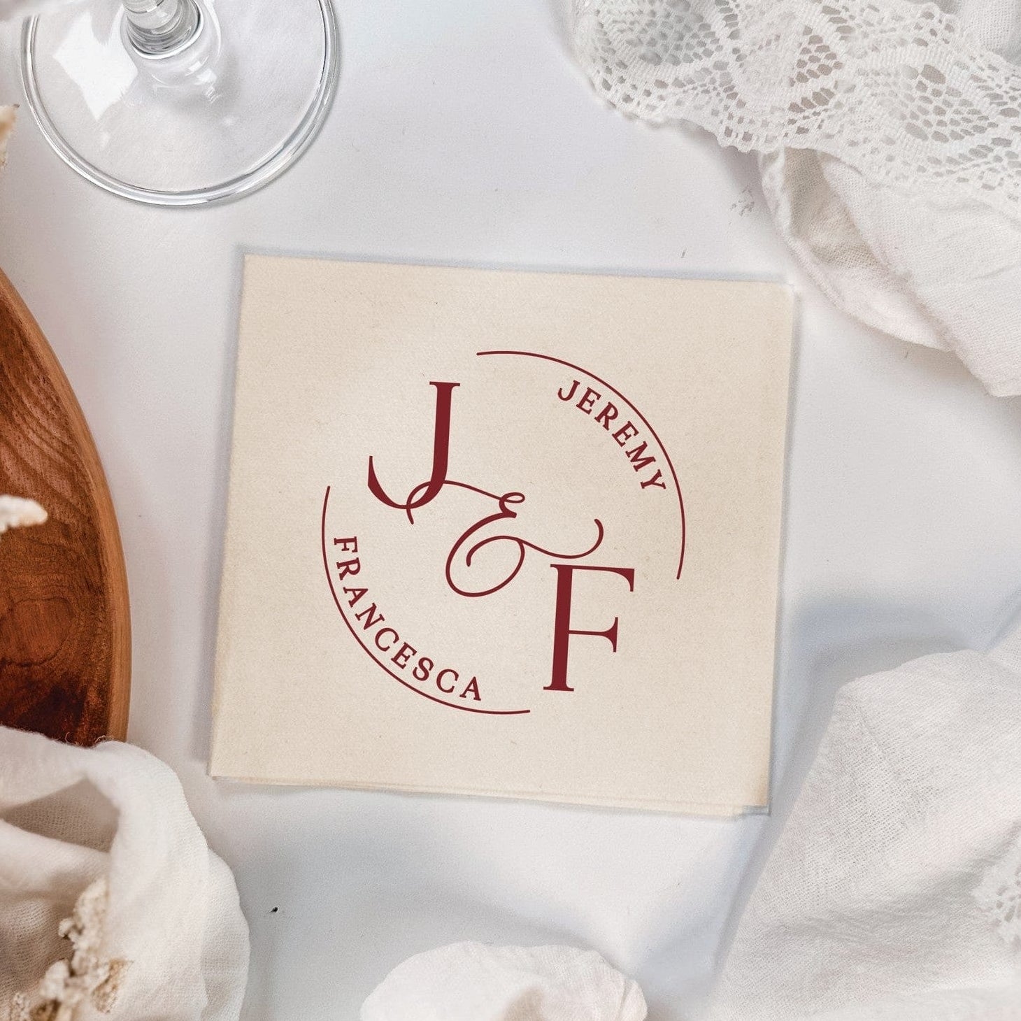 An off-white square linen cocktail napkin features a custom deep red circular monogram design, perfect for a wedding or elegant event.