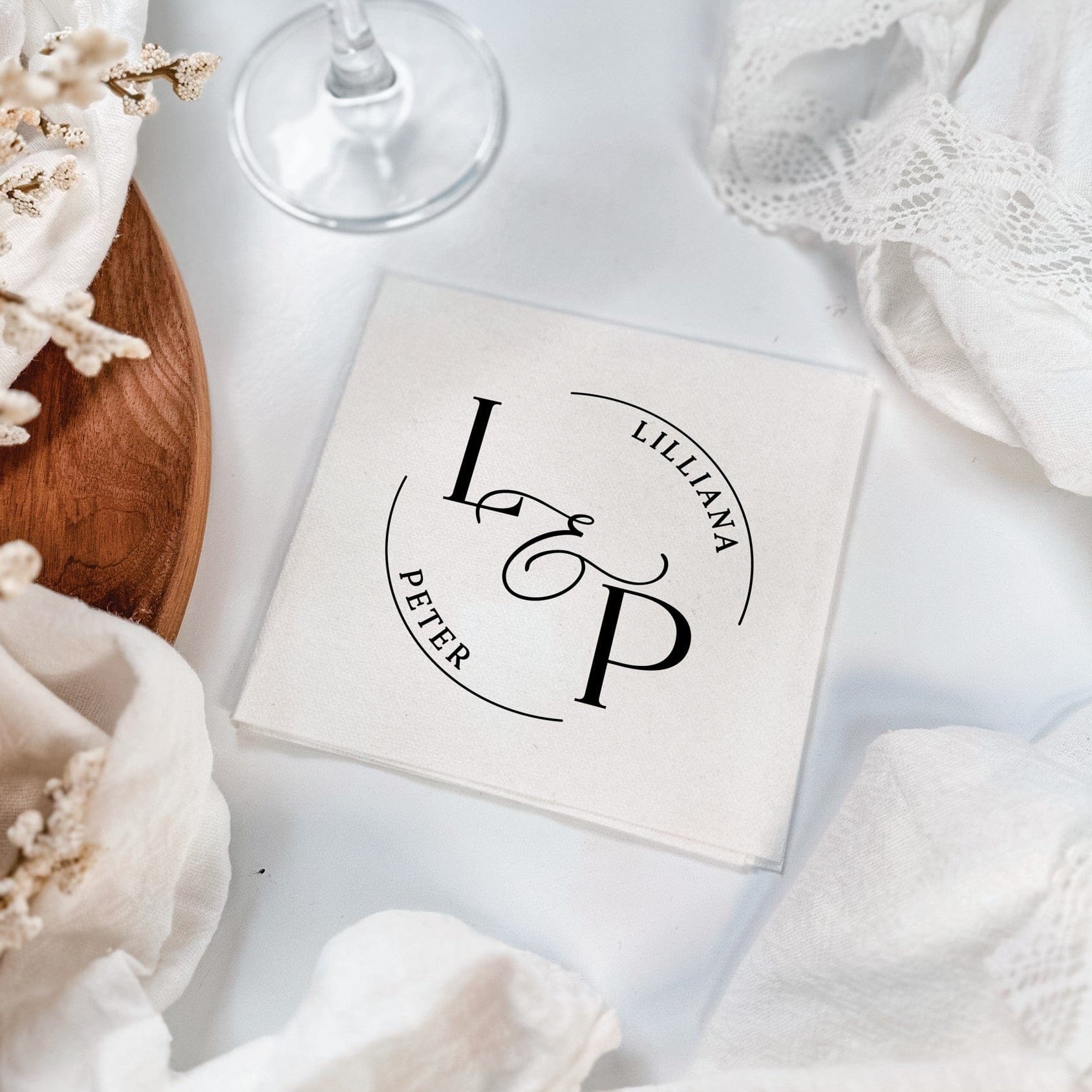A square white paper napkin features a custom black circular monogram design, perfect for a wedding or elegant event.