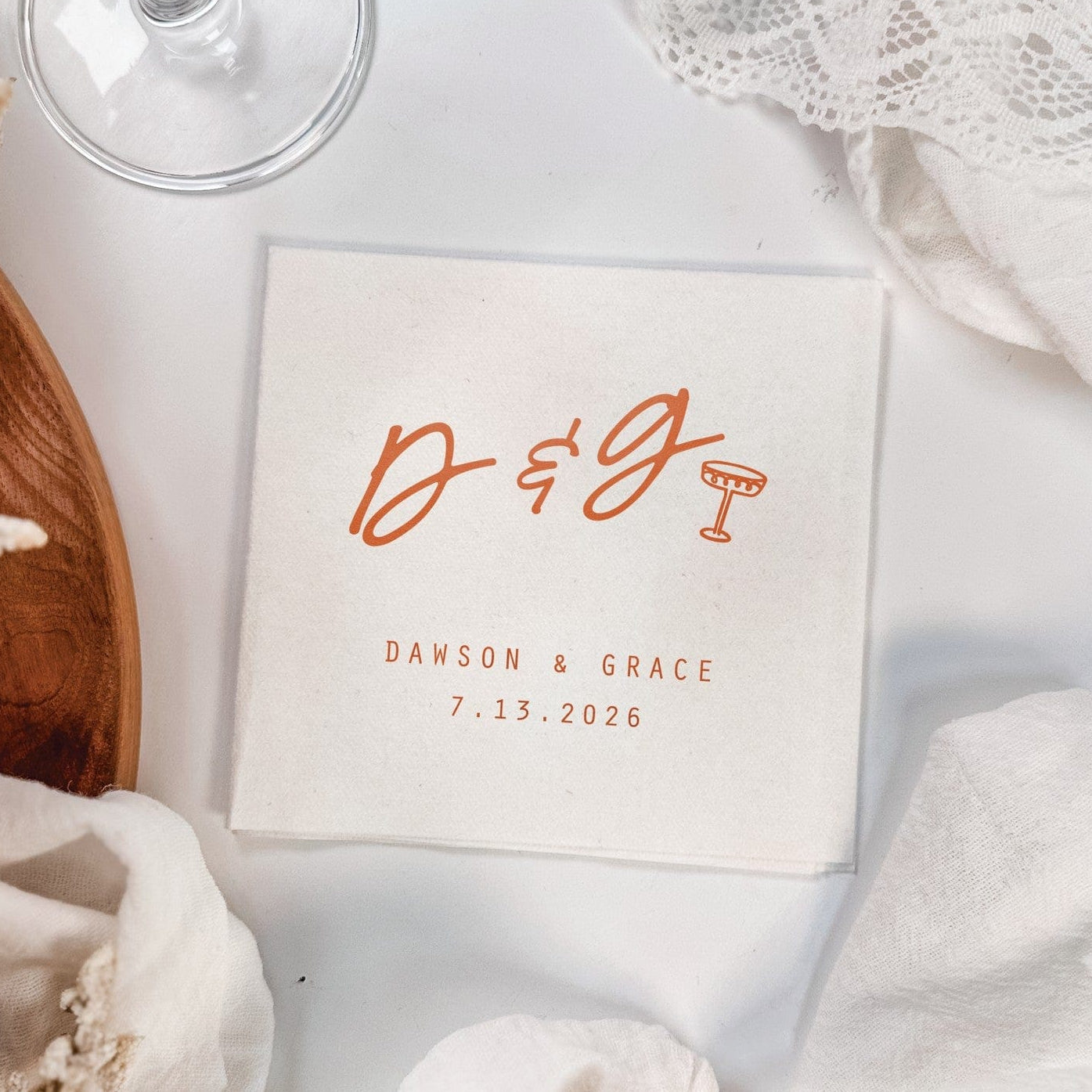 A square, light-colored personalized wedding or event napkin features custom initial calligraphy and a small cocktail glass icon printed in orange, placed on a white surface with draped decorative fabric.