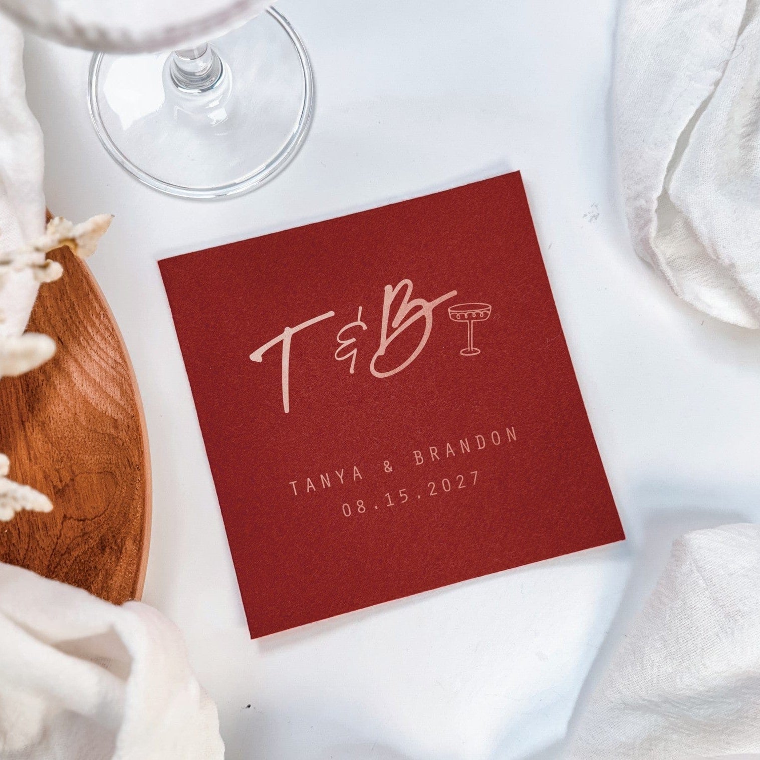 A square, personalized red paper cocktail napkin featuring custom initial typography and a champagne glass icon, perfect for a wedding or special event.