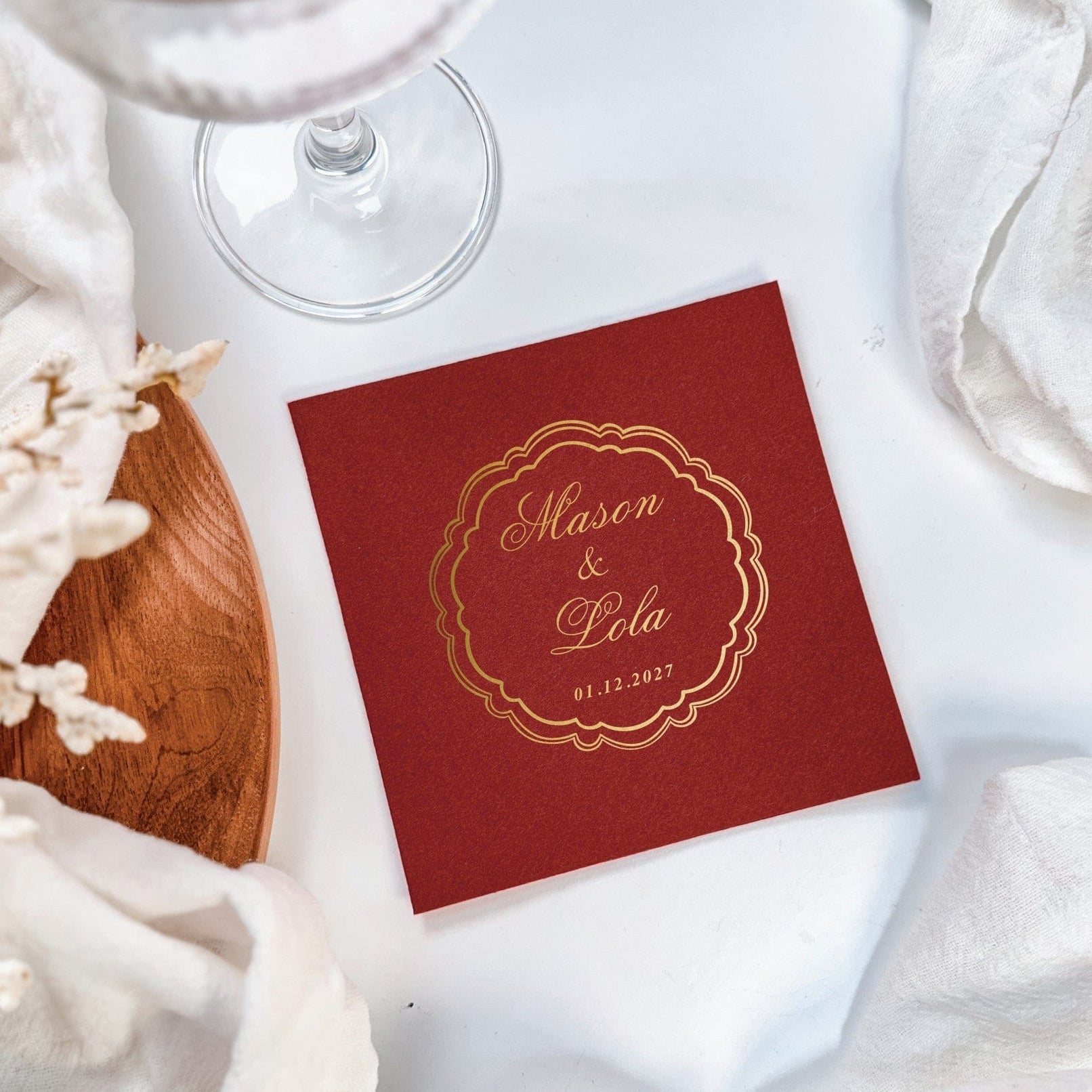 A square deep red personalized linen-feel napkin features a metallic gold foil scrollwork frame design, ideal for a wedding or elegant event.
