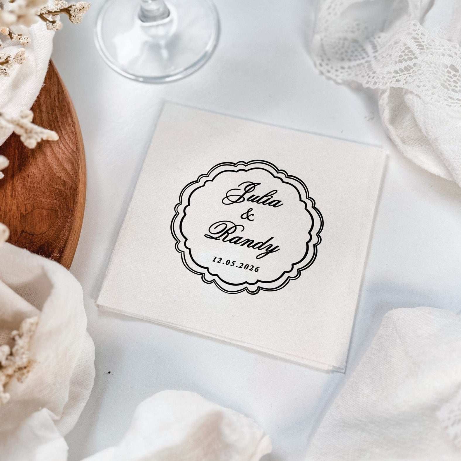 A stack of white linen-feel napkins featuring a black printed scalloped frame design, presented with white fabric and dried flowers, perfect for elegant wedding event decor.