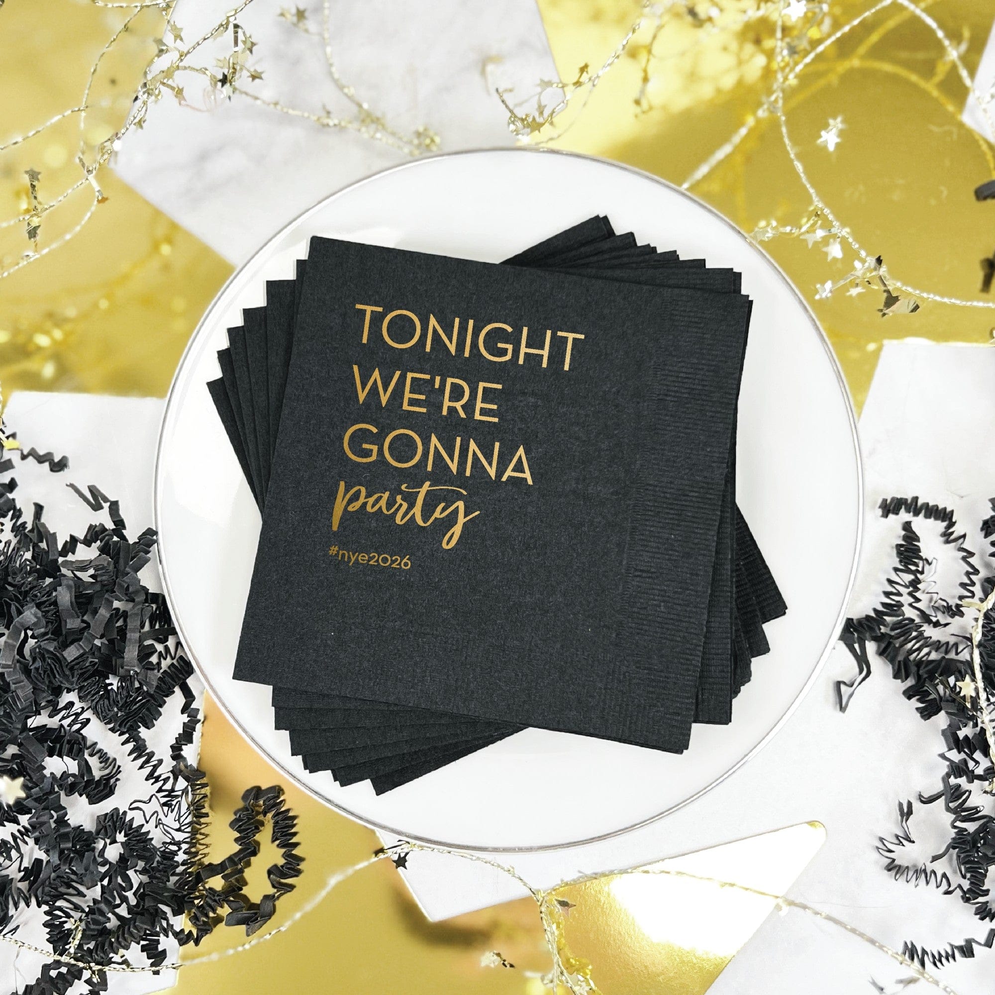 A stack of black paper cocktail napkins with elegant gold foil lettering is arranged on a white plate amidst black shredded paper and gold star garland, ideal for a festive celebration.