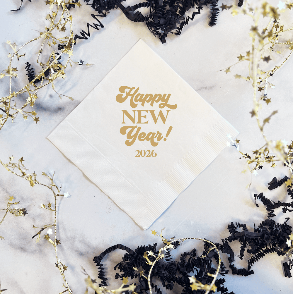 White napkin with gold foil stamped text reading "Happy New Year!" and "2026" surrounded by gold and black party decorations.