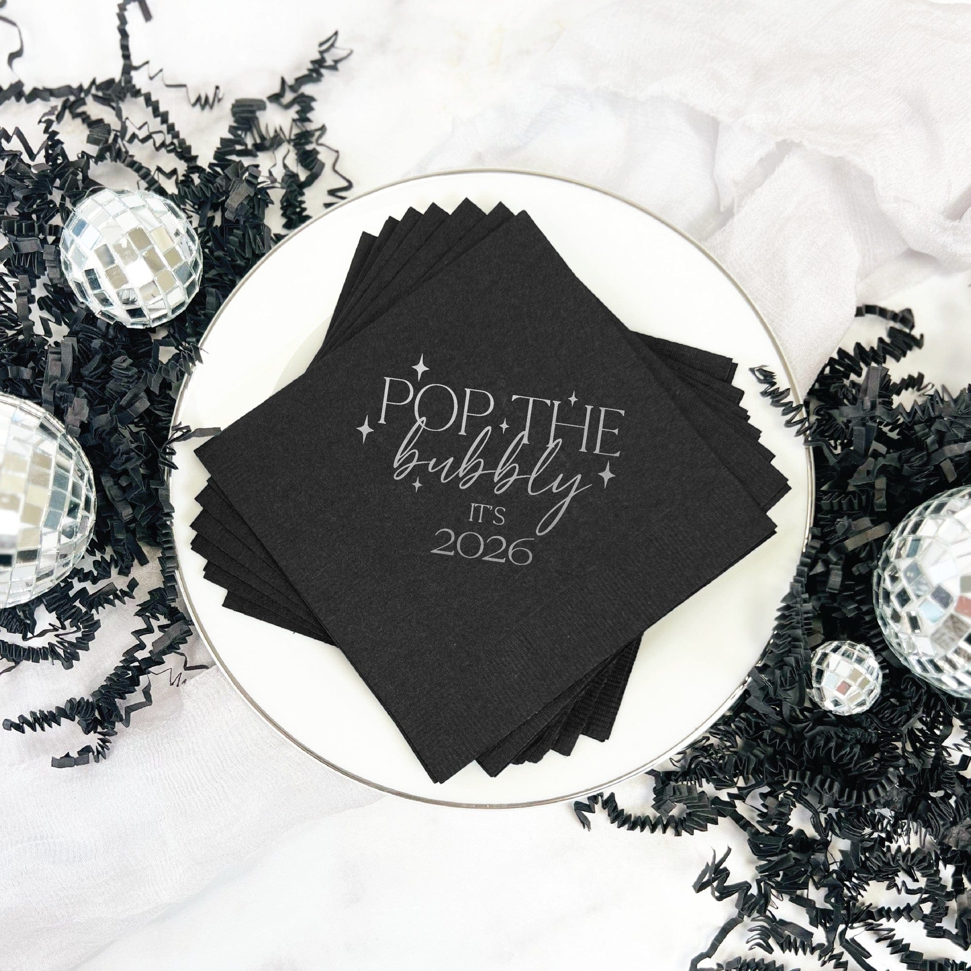 A stack of black cocktail napkins featuring a metallic silver celebratory design with stars and mixed typography is presented on a white plate, ideal for a festive party or event.