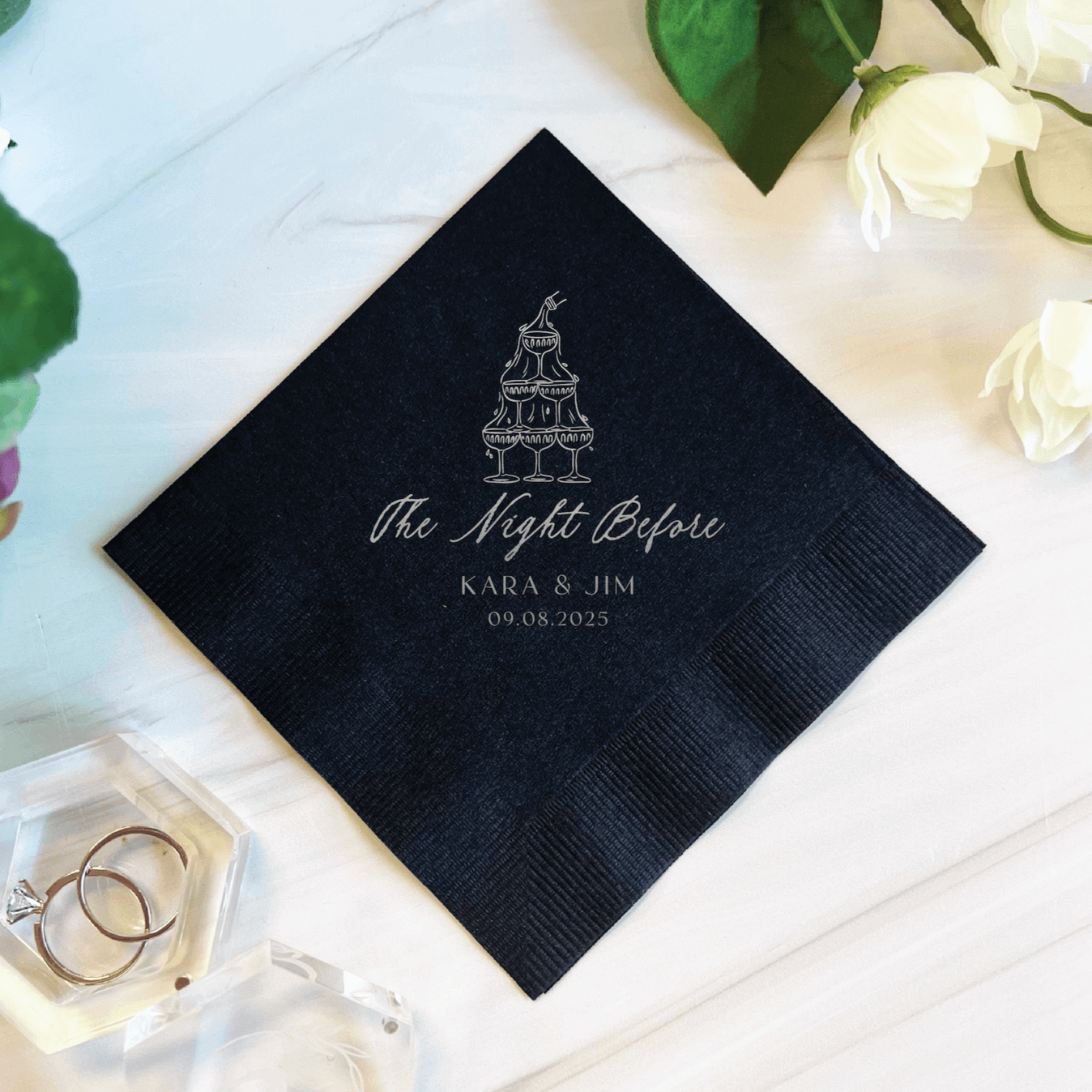 Black cocktail napkin with silver foil stamped wedding cake design and "The Night Before" text with couple's names and date.