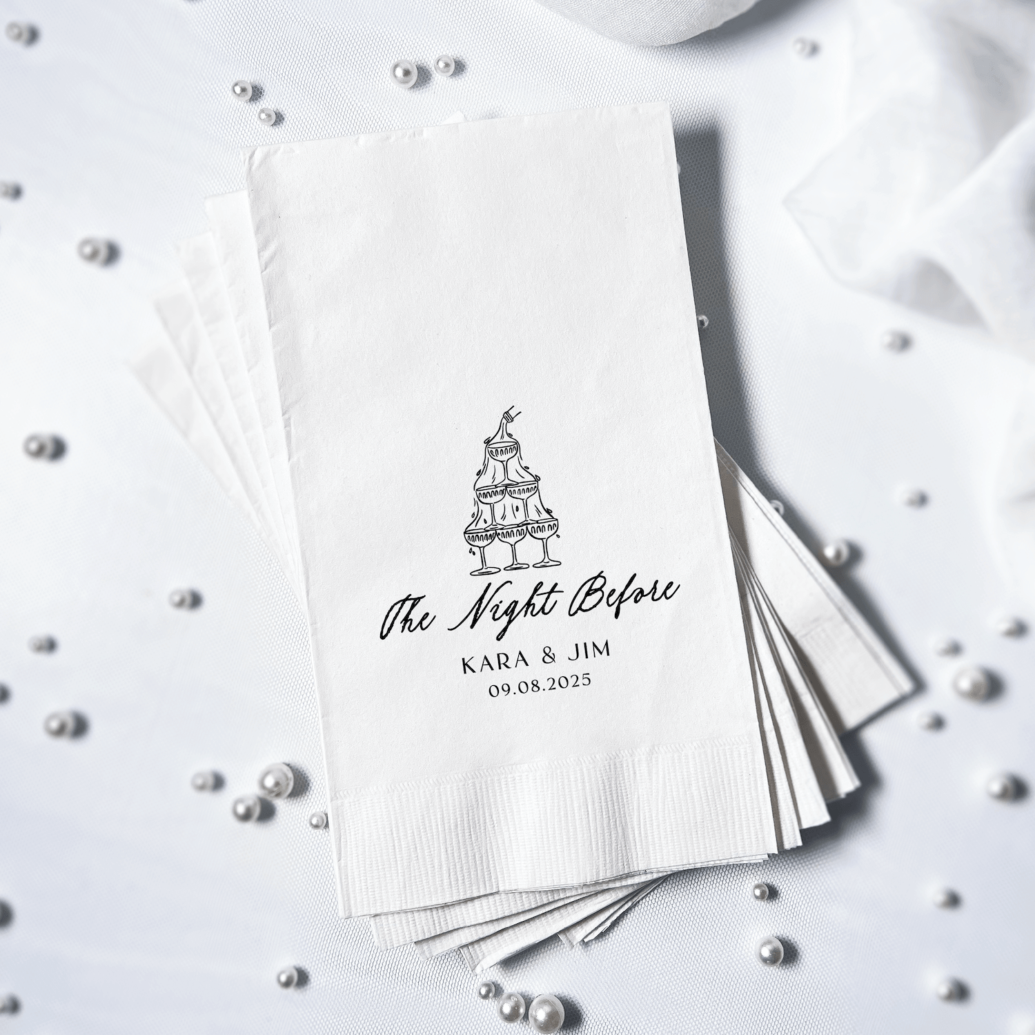 Stack of white napkins with black foil-stamped wedding cake design and "the Night Before" text personalized with couple's names and date.