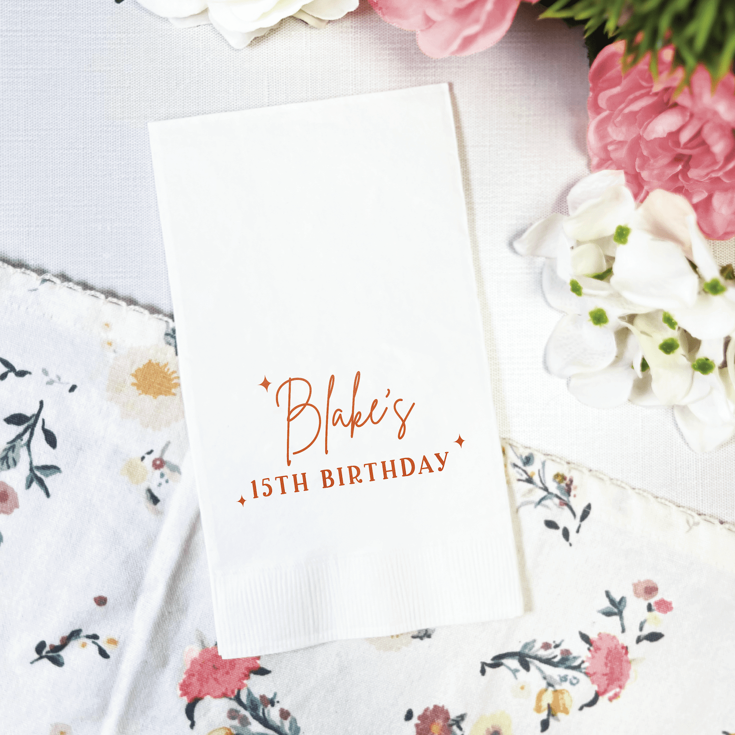 White napkin with rose gold foil stamped text reading "Blake's 15th Birthday" surrounded by pink and white flowers