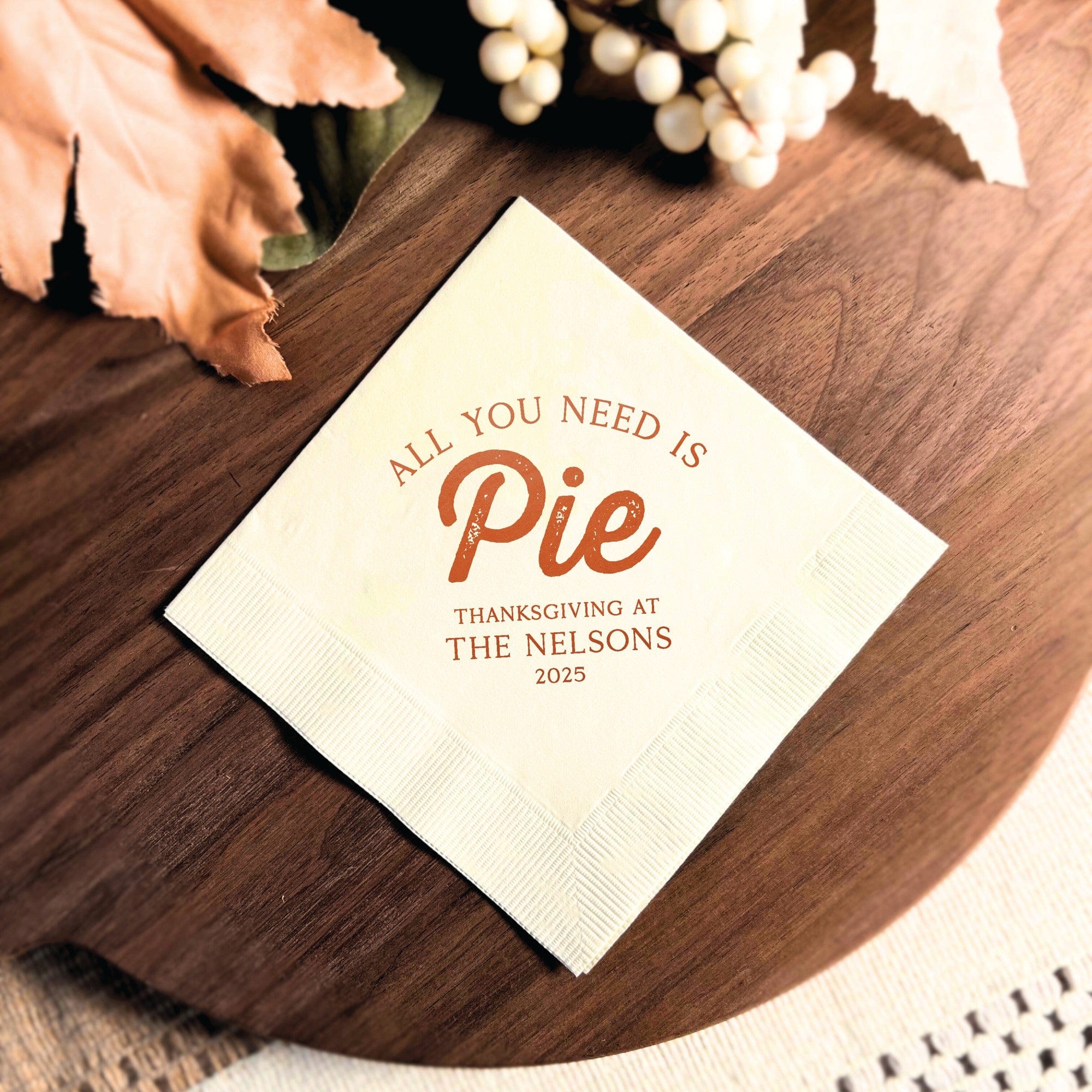 A square, cream-colored paper napkin featuring a textured border and custom burnt orange typography, designed for a Thanksgiving celebration.